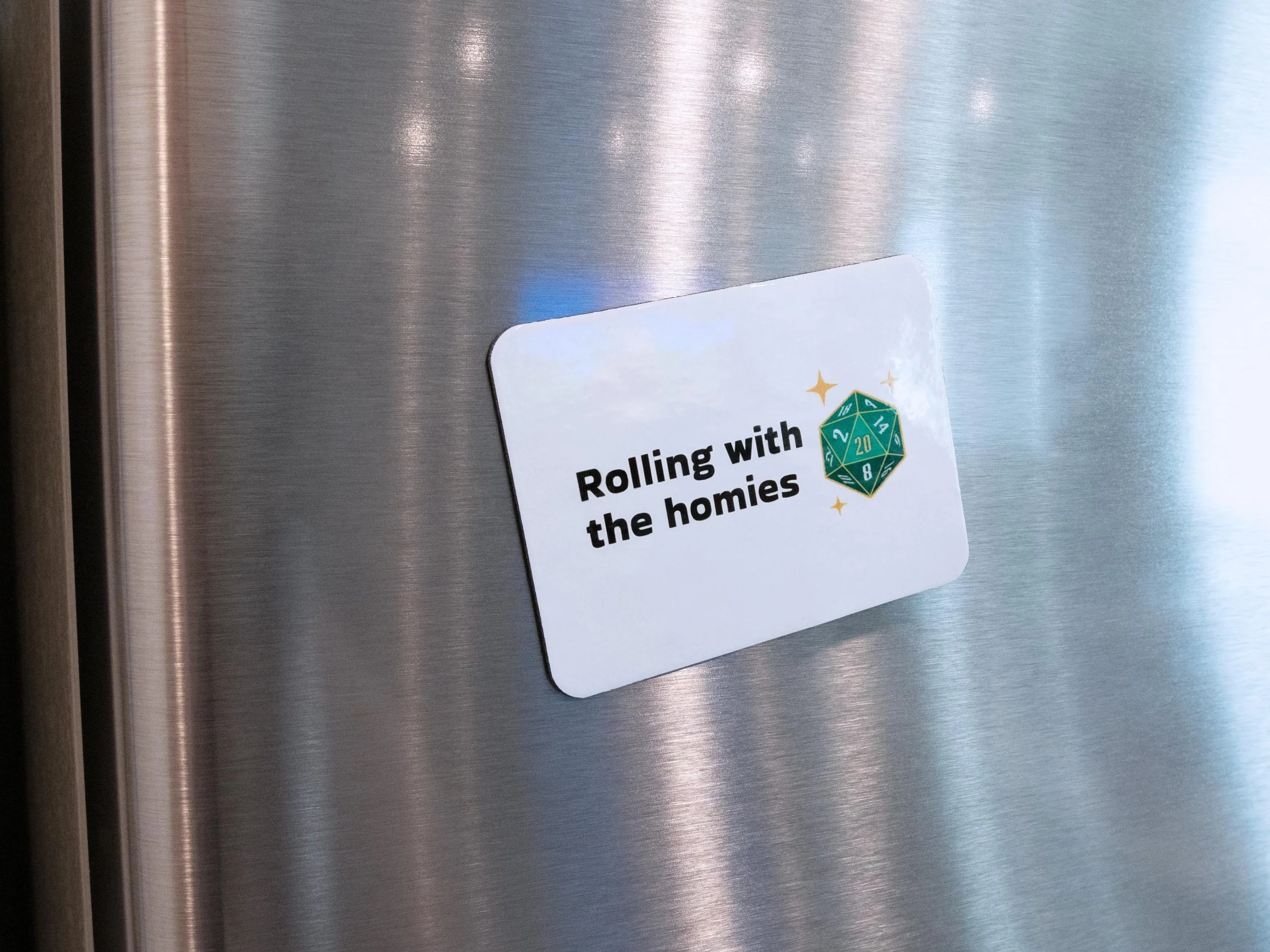 A magnet on a stainless steel fridge. The design is a green d20 die with text that says “Rolling with the homies”