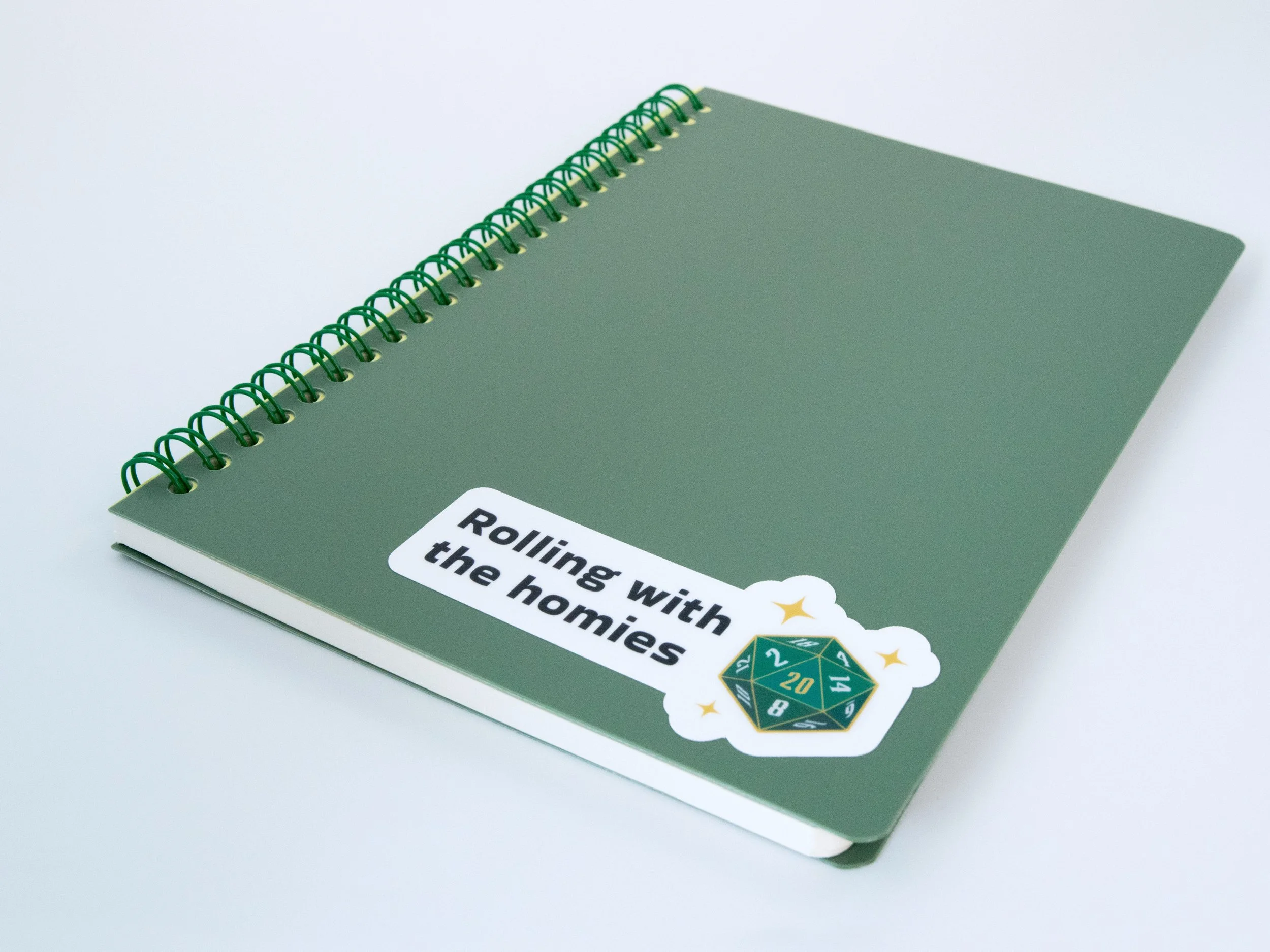 A sticker of a green d20 die with text that says “Rolling with the homies”. The sticker is on a green notebook