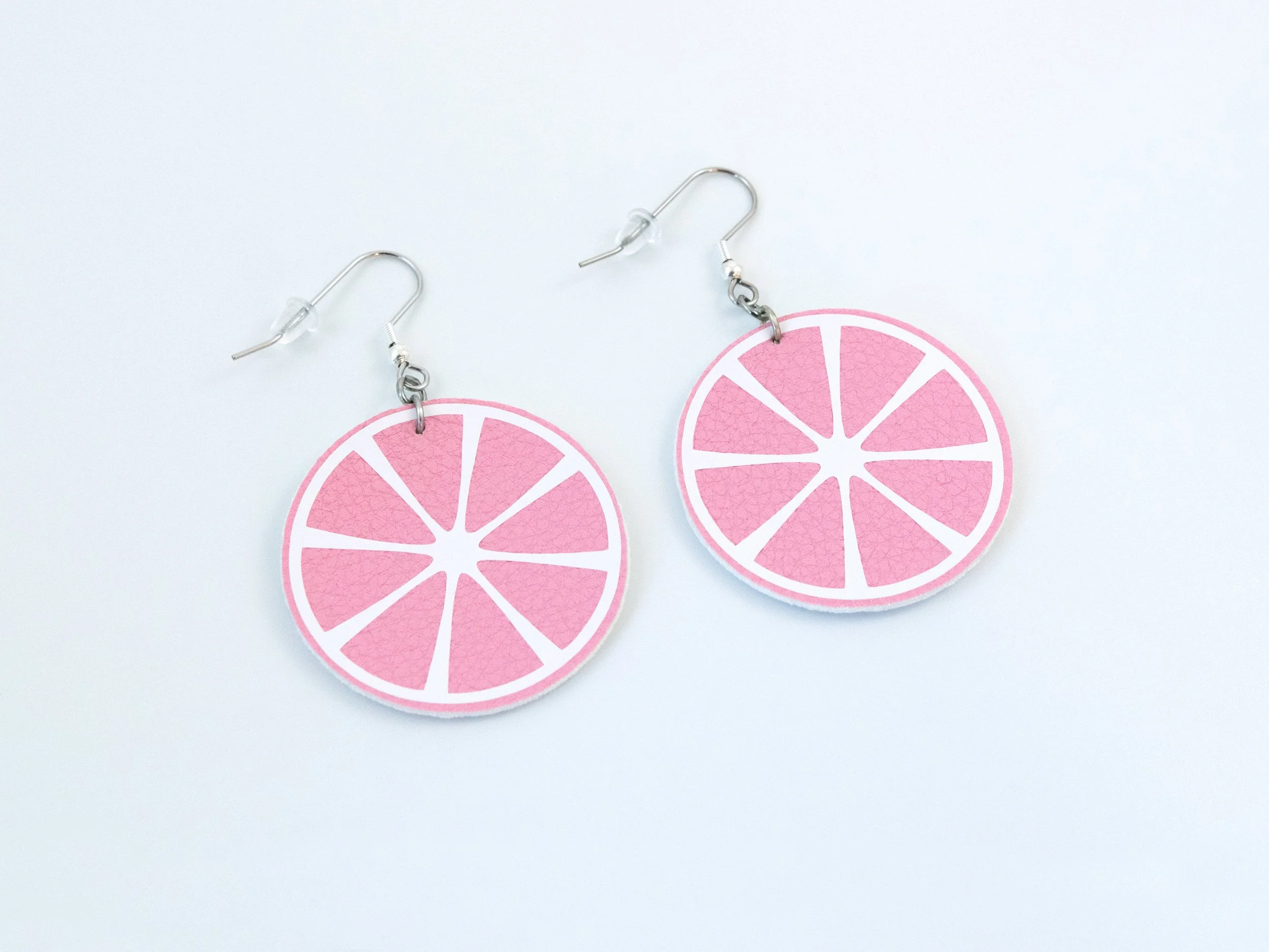 Angled view of round citrus slice earrings. They have pink faux leather, white vinyl details, and silver-finished hooks