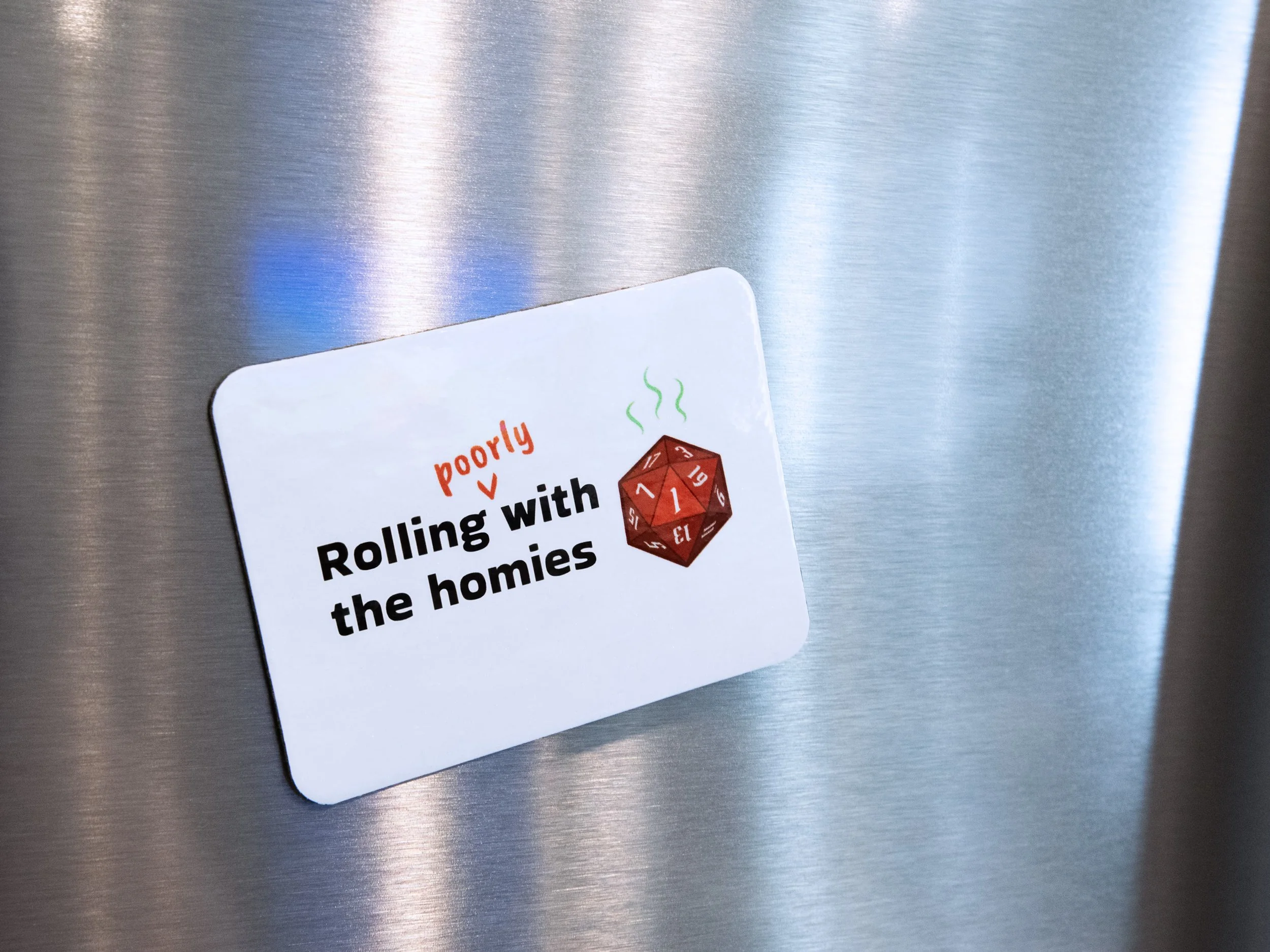 A magnet on a stainless steel fridge. The design is a red d20 die with text that says “Rolling poorly with the homies”