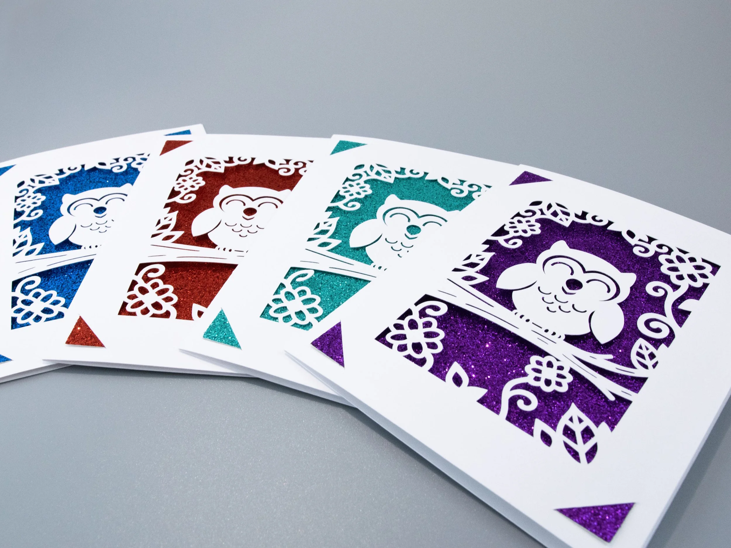 Angled view of 4 owl-themed greeting cards fanned out across a table. Each card has a different glitter background color