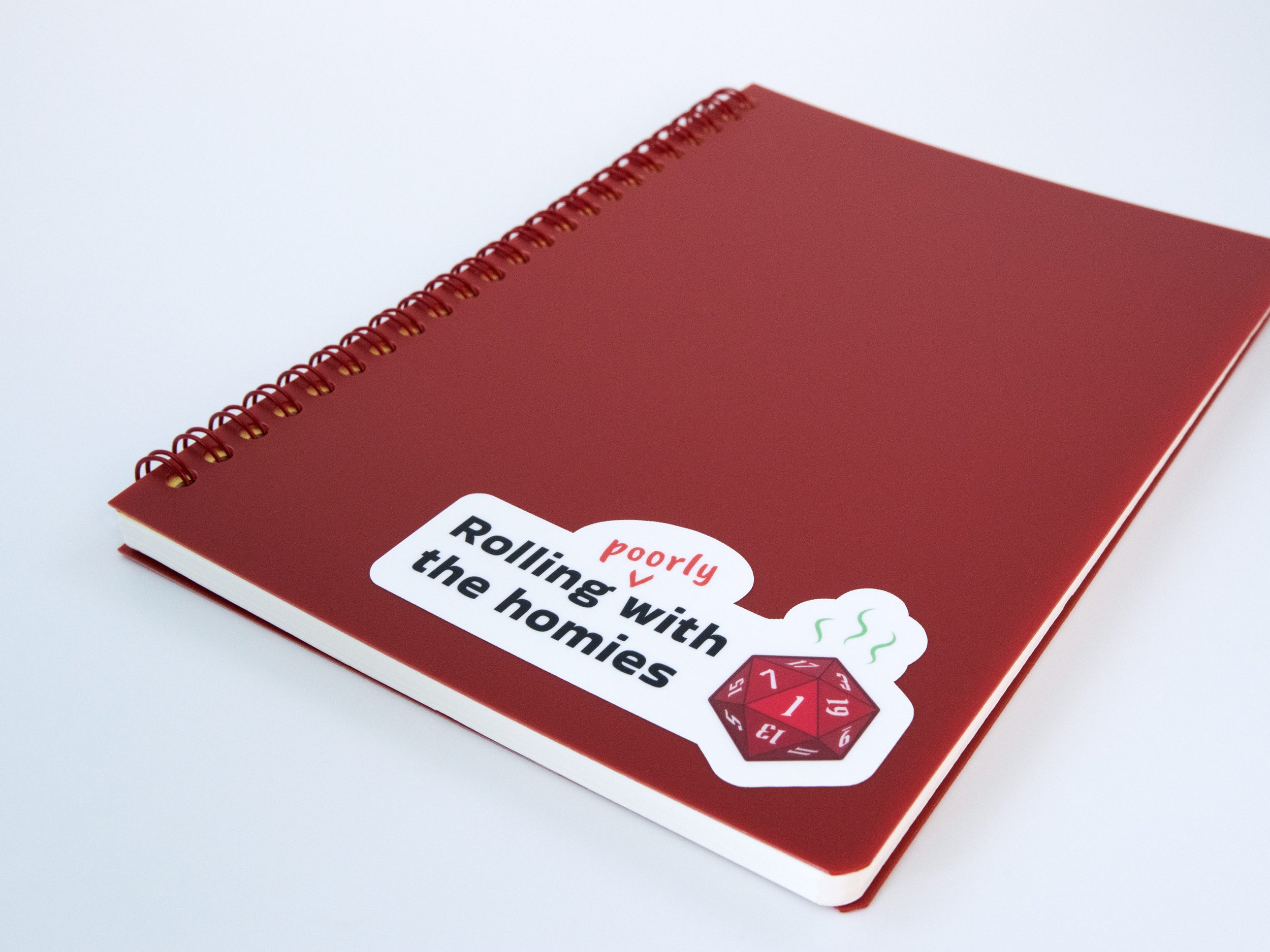 A sticker of a red d20 die with text that says “Rolling poorly with the homies”. The sticker is on a red notebook
