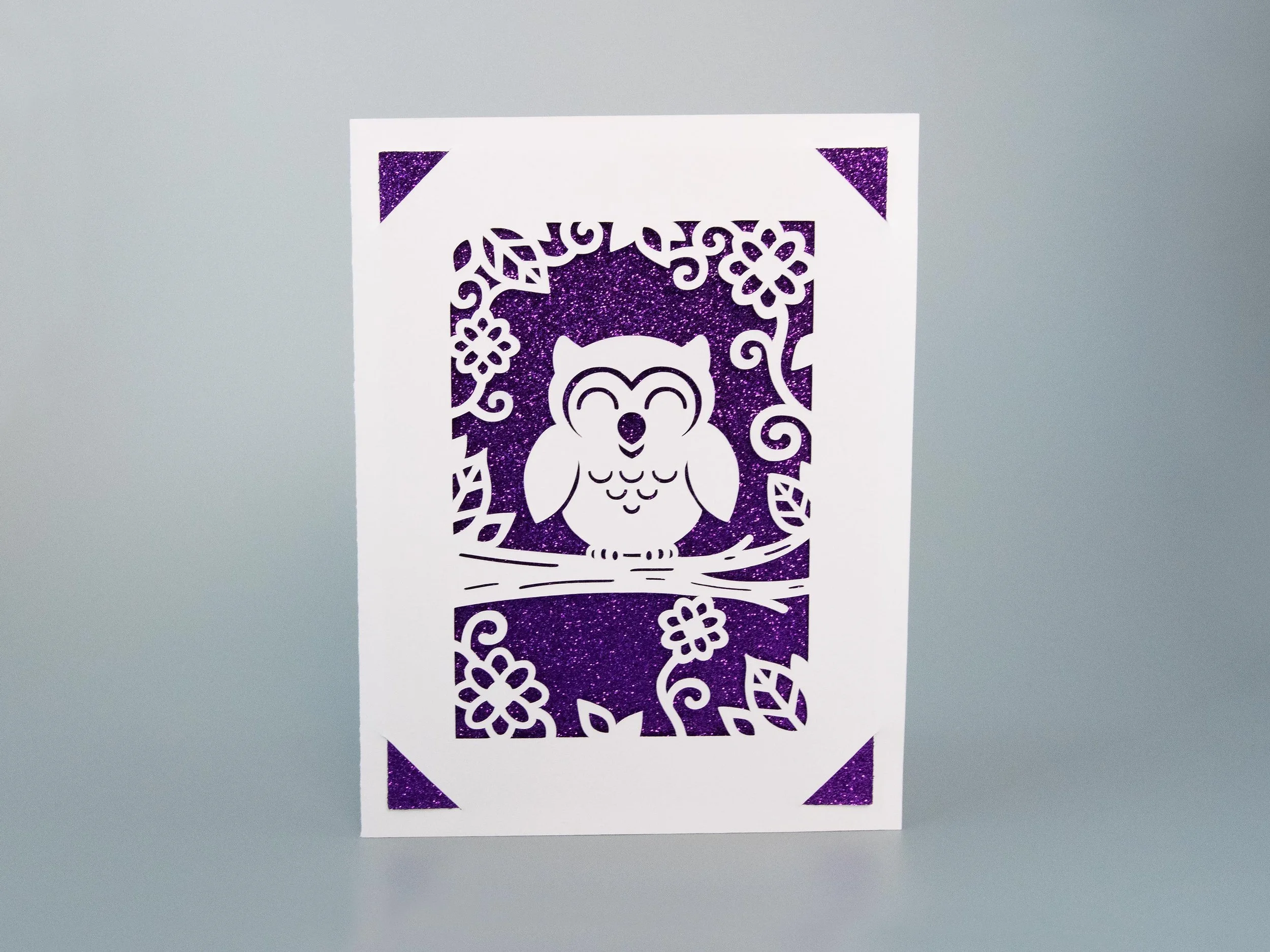 Front view of a greeting card with an owl cutout and purple glitter background