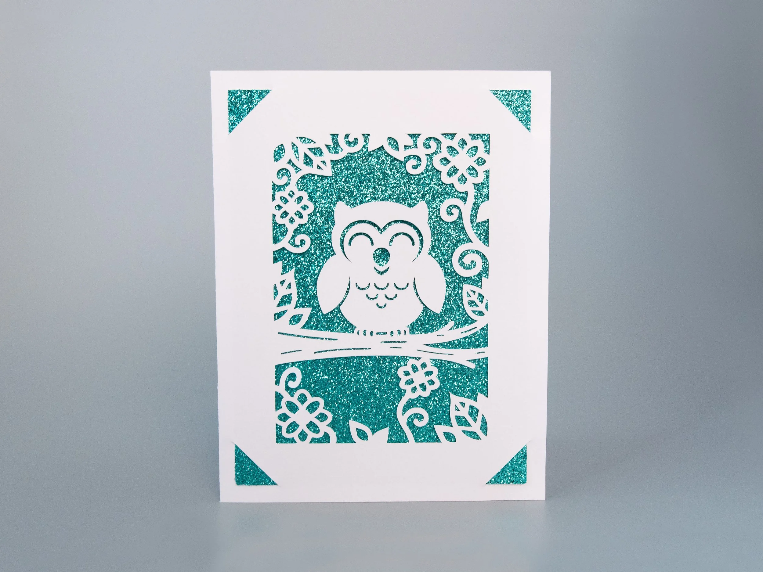 Front view of a greeting card with an owl cutout and turquoise glitter background
