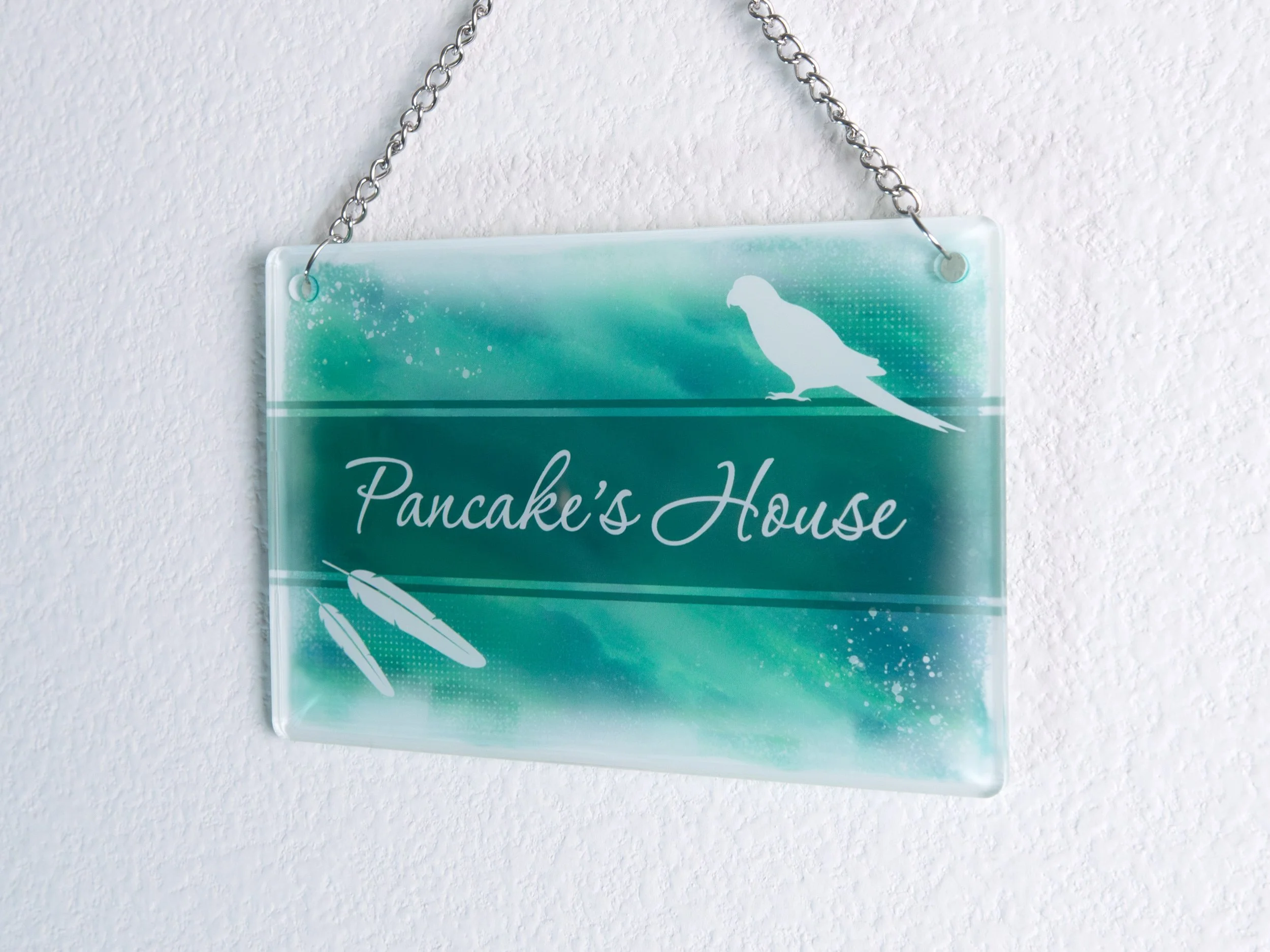 A small acrylic sign, which is hung up on the wall by a metal chain. It has bird-themed artwork with text that says "Pancake’s House"