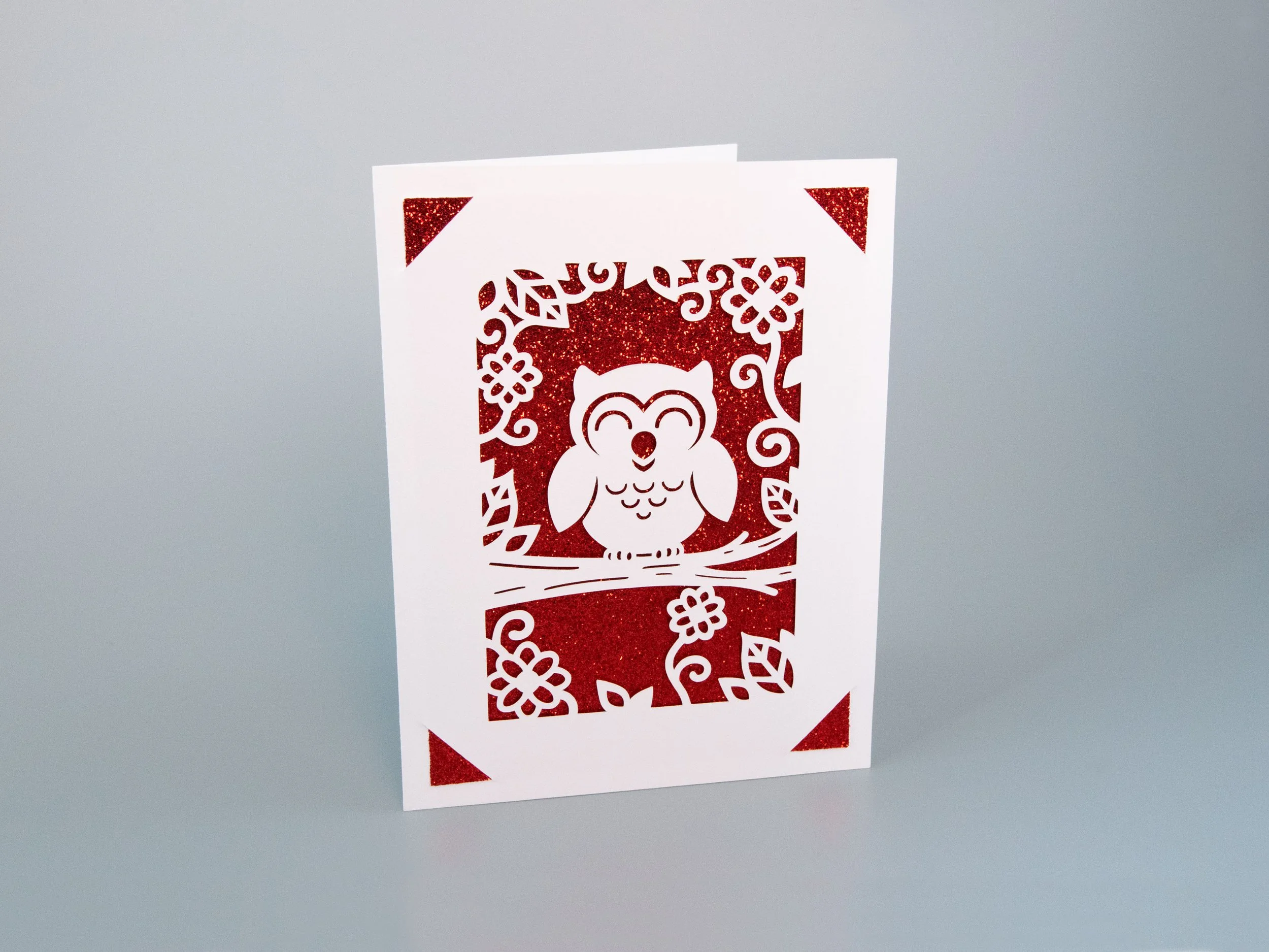 Angled view of a greeting card with an owl cutout and red glitter background