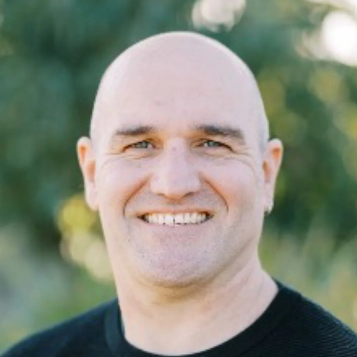 Close-up of a smiling man with a shaved head, outdoors with blurred green trees in the background.