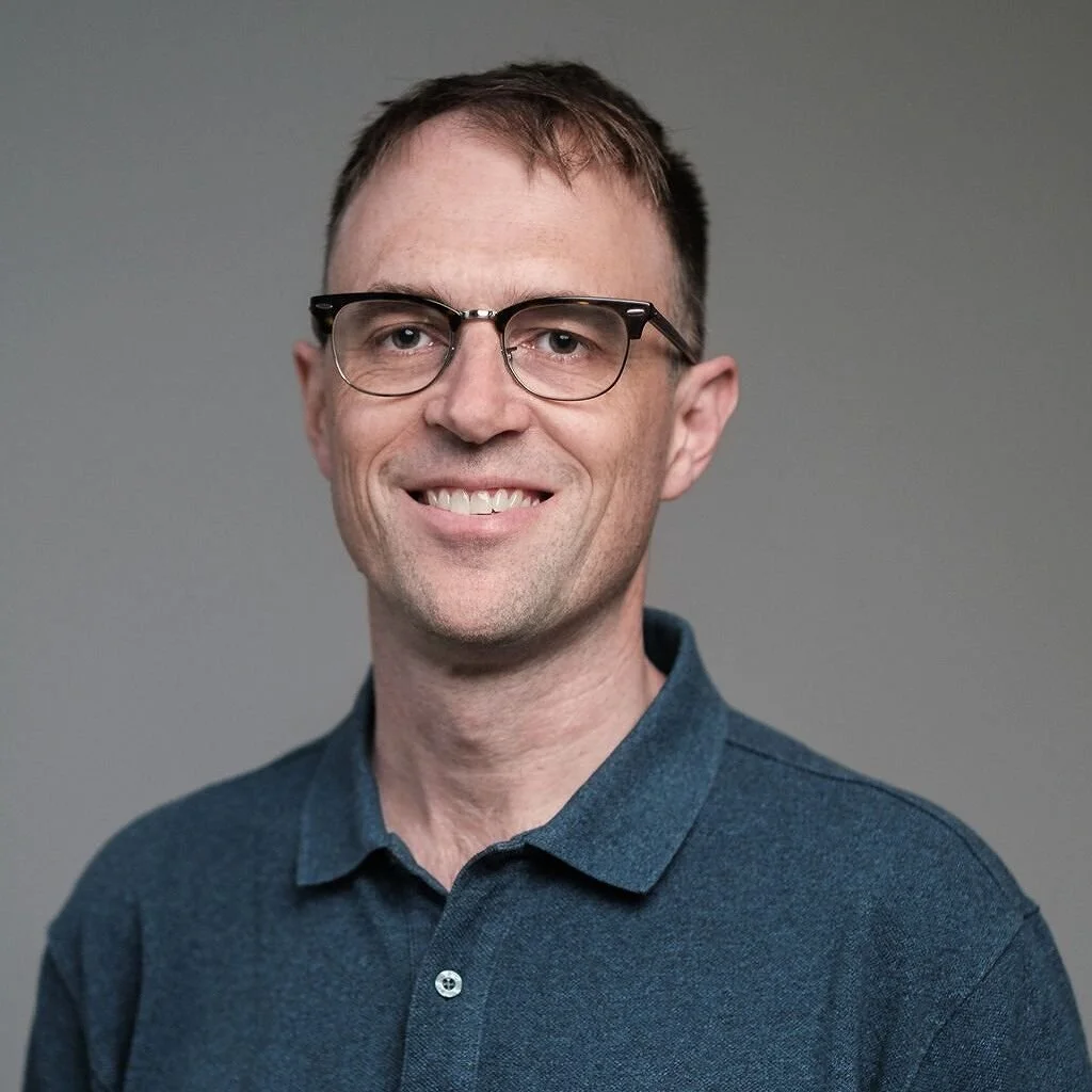 Headshot of a smiling man wearing glasses and a dark blue collared shirt against a gray background.
