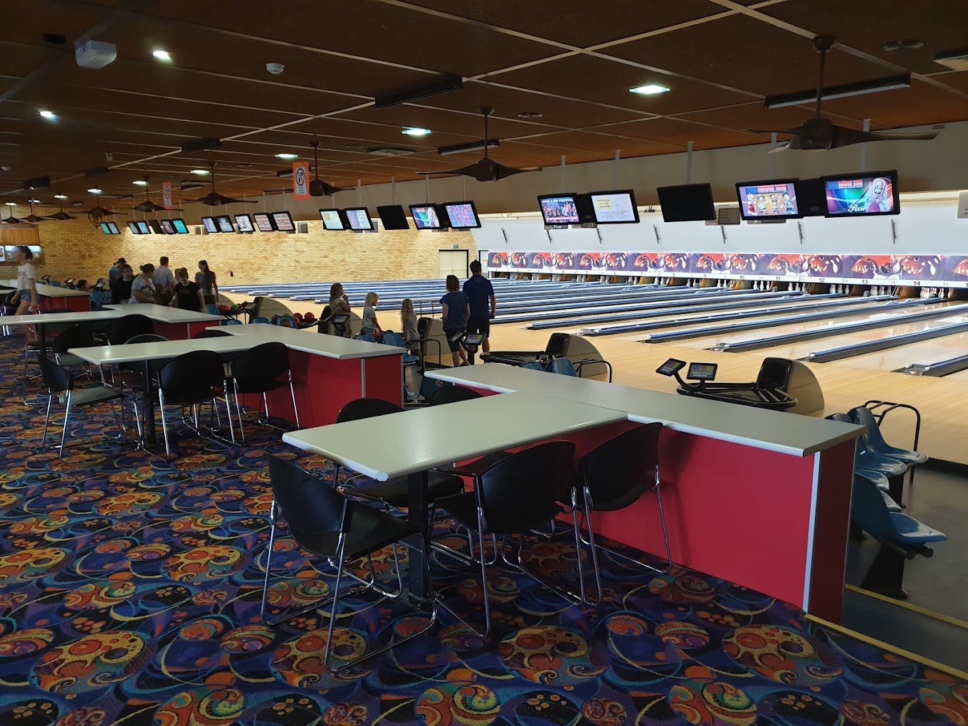 A bowling alley with several lanes, digital scoreboards above, and social groups of people playing and observing. There are tables and chairs in the foreground and a colorful carpet with abstract patterns.
