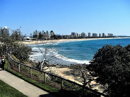 Scenic view of a beach with ocean waves, a sandy shoreline, and city buildings in the background. Trees and a walkway with a person walking are in the foreground.