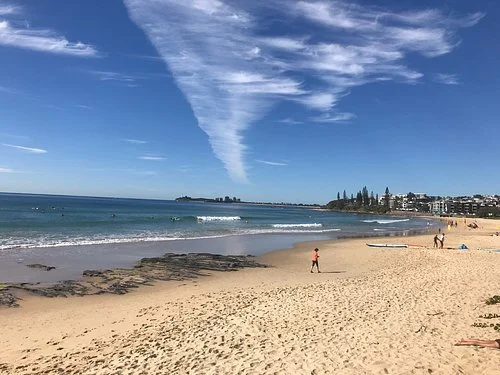 Sunny beach with people walking along the shoreline, a rocky area on the sand, and buildings in the distance under a blue sky with clouds.