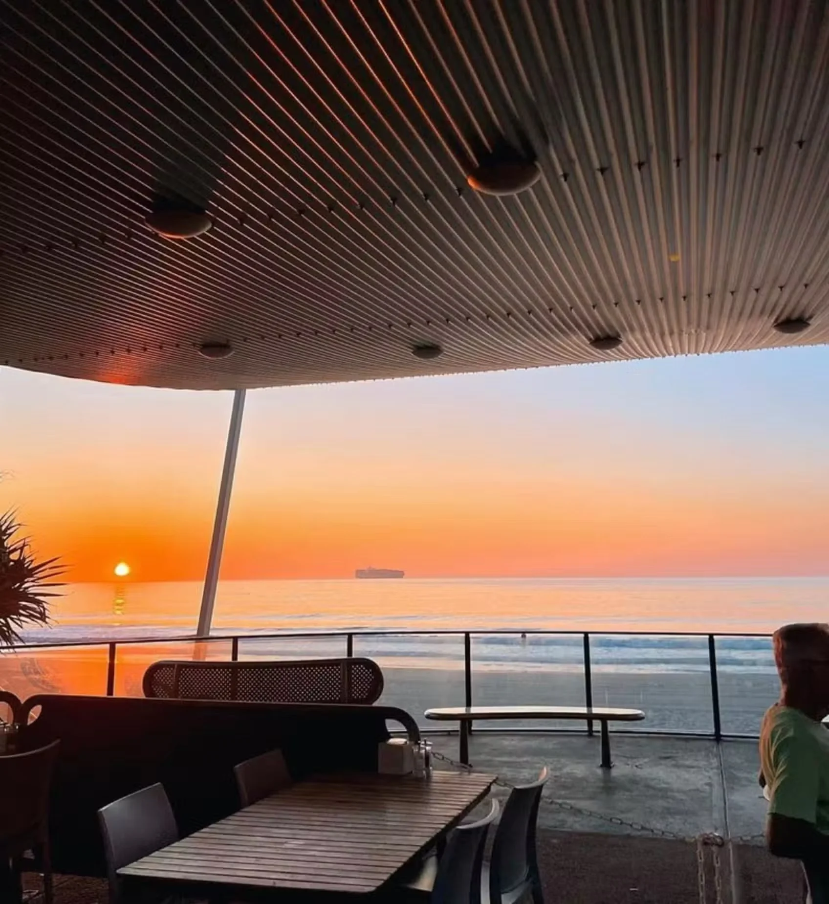 Beachside seating area with tables and chairs under a canopy, overlooking the ocean at sunset, with a ship in the distance and a person standing near the railing.