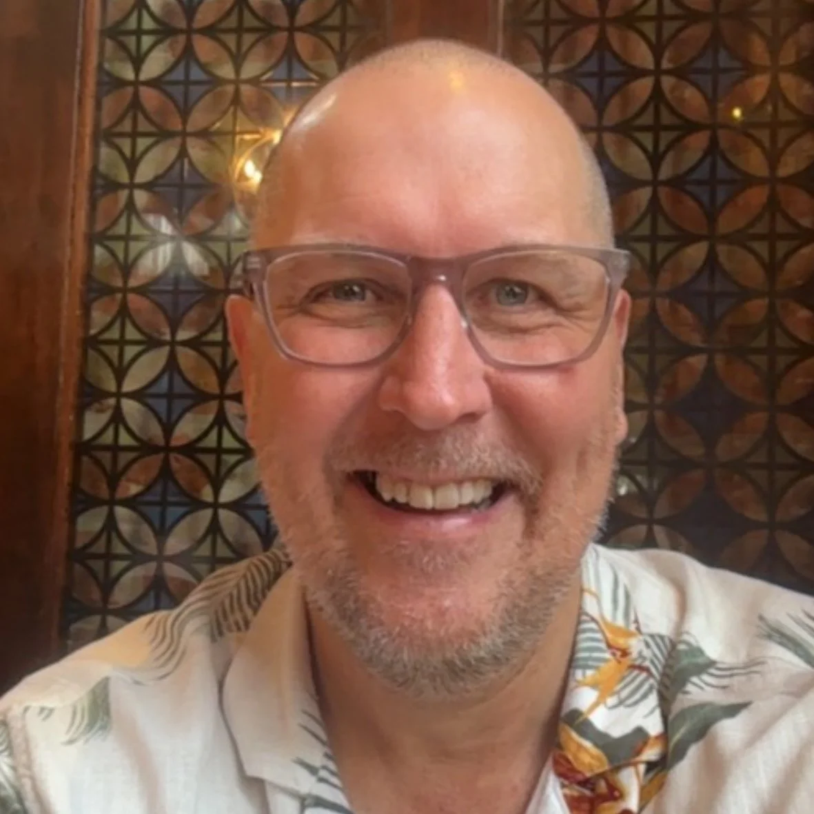 A smiling middle-aged man with glasses, a beard, and a tropical-patterned shirt, in front of an intricate wooden lattice background.