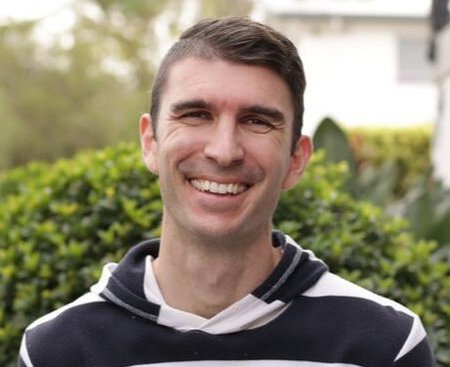 A smiling man outdoors with greenery and a house in the background.