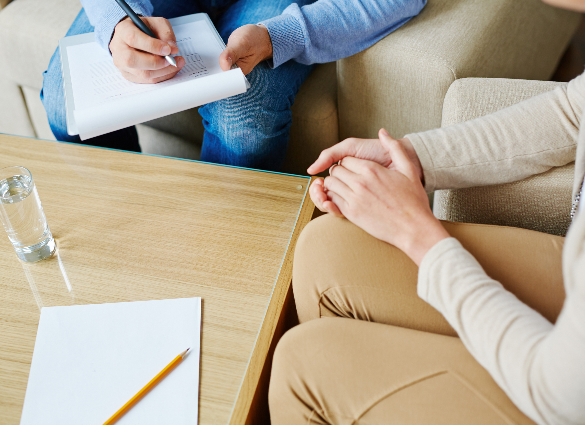 Two people sitting at a wooden table during a consultation; one person taking notes with a pen, the other has hands clasped. There is a glass of water and a blank sheet of paper with a pencil on the table.