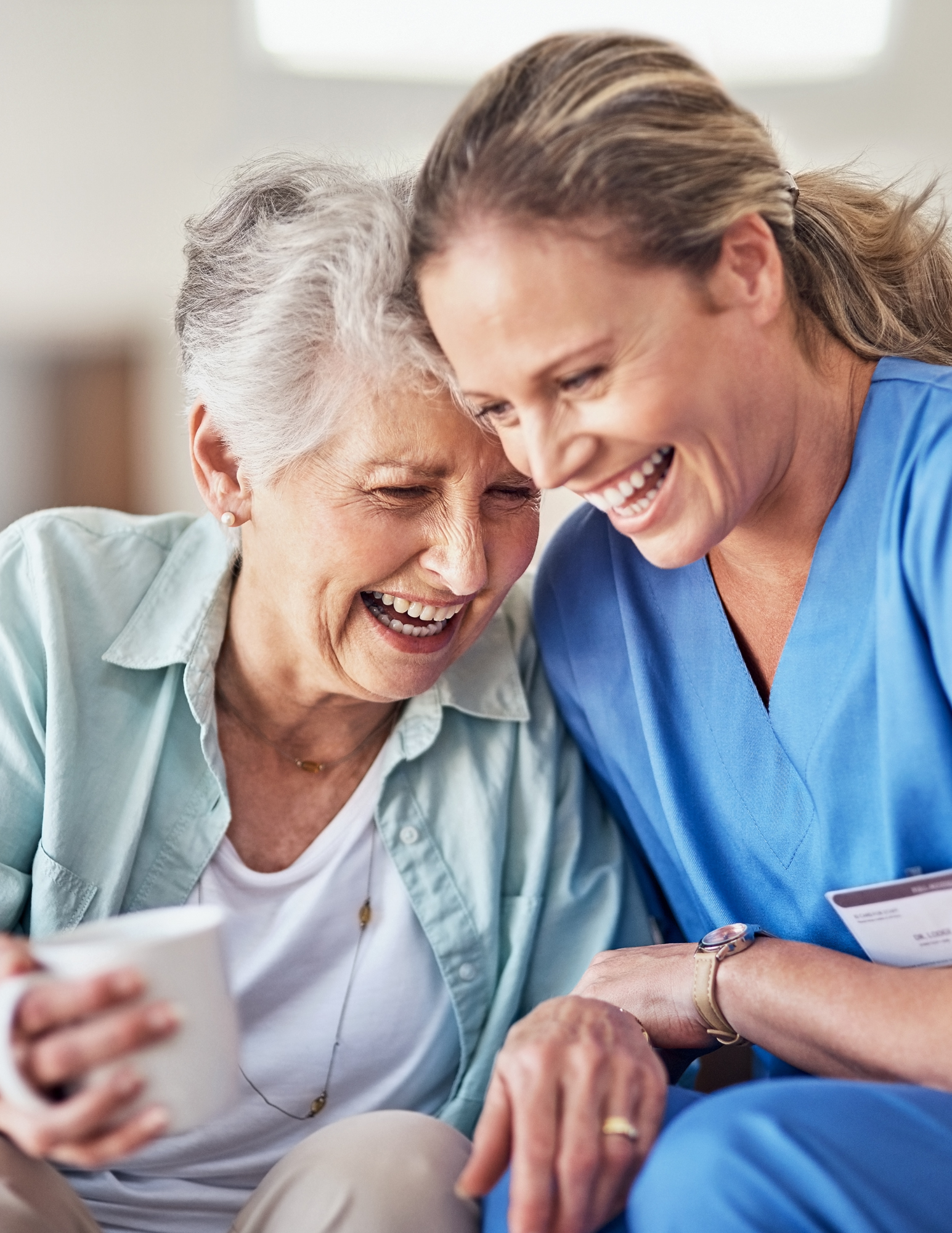 An elderly woman and a healthcare worker sharing a joyful moment, both smiling and leaning towards each other.
