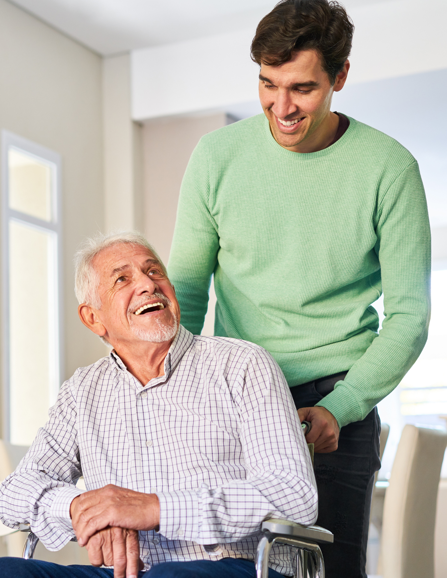 A young man smiling and helping an elderly man in a wheelchair, both indoors in a bright room.