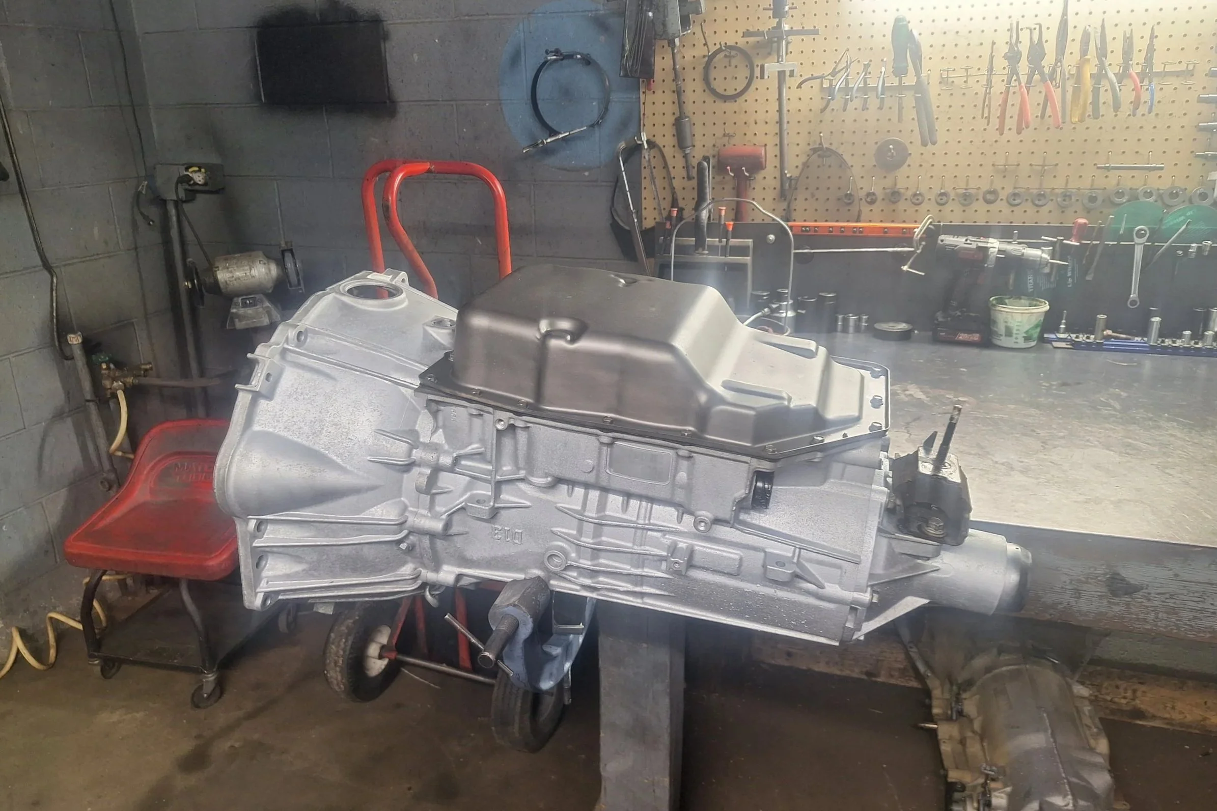 A car transmission sits on a workbench in a garage or workshop, with tools hanging on a pegboard and various equipment in the background.