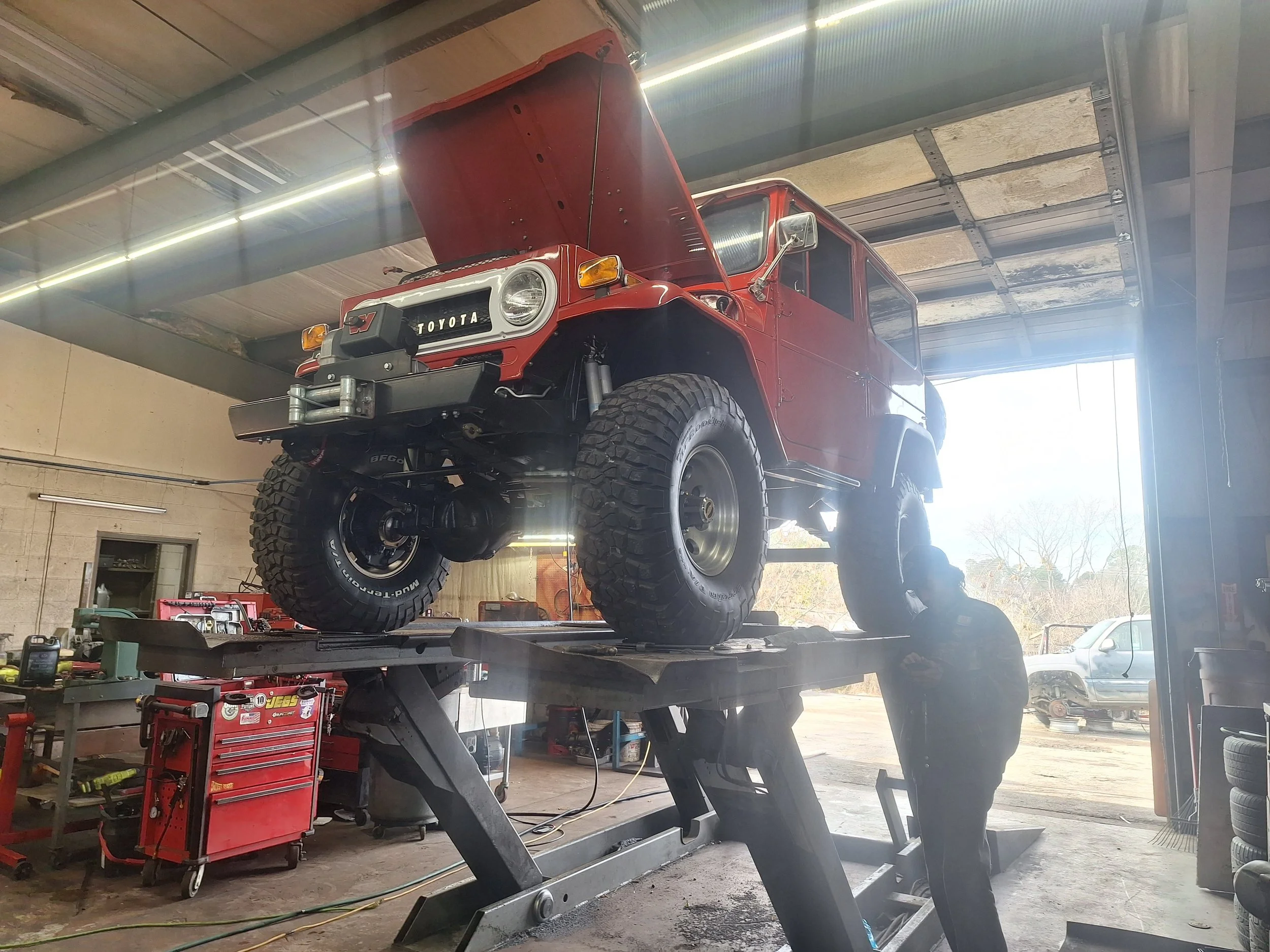 A red vintage Toyota Land Cruiser on a hydraulic lift inside an auto repair shop, with a mechanic working underneath.