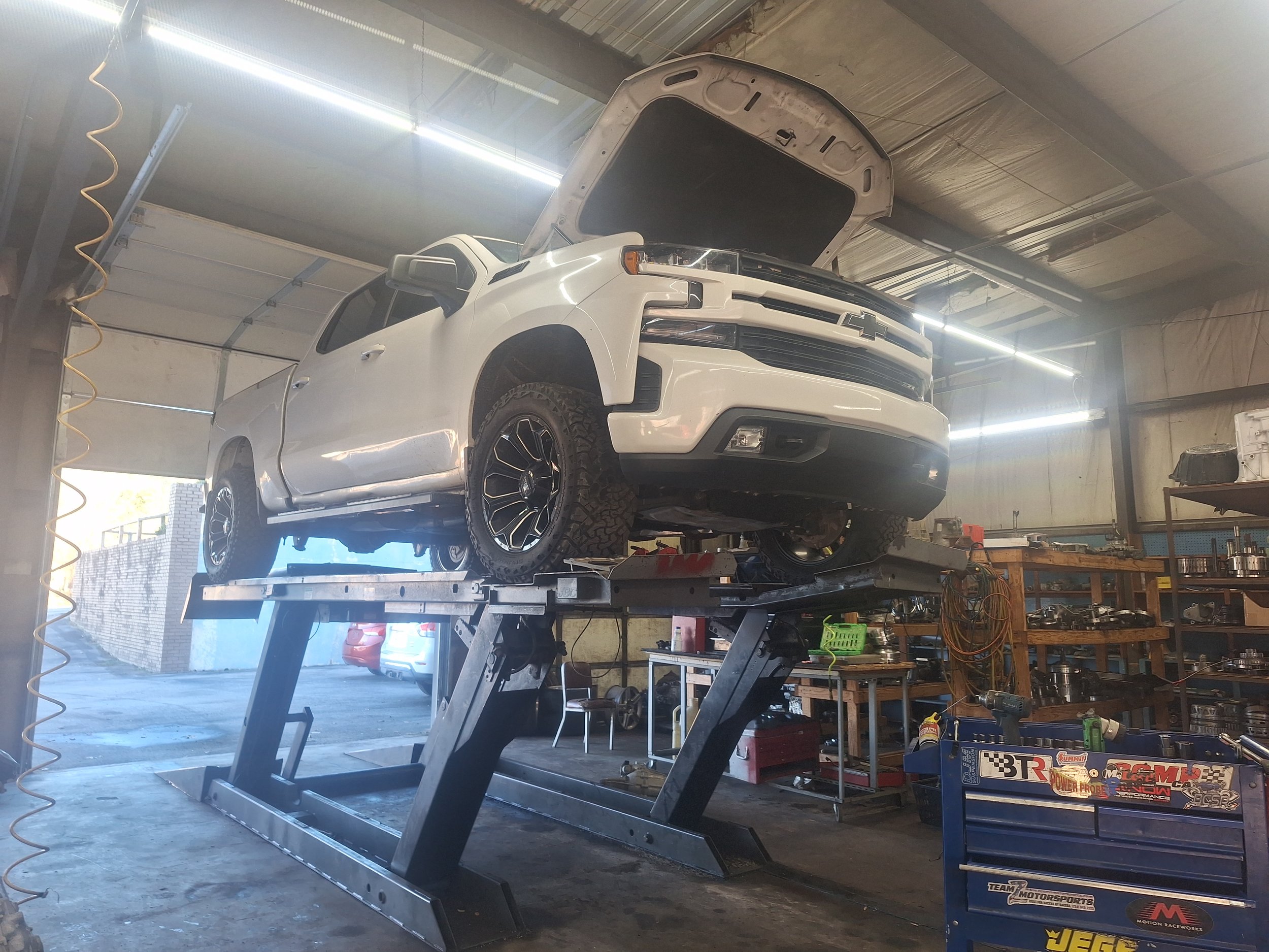 A white Chevrolet pickup truck raised on a hydraulic lift inside an automotive repair shop, with the hood open for maintenance or repair.