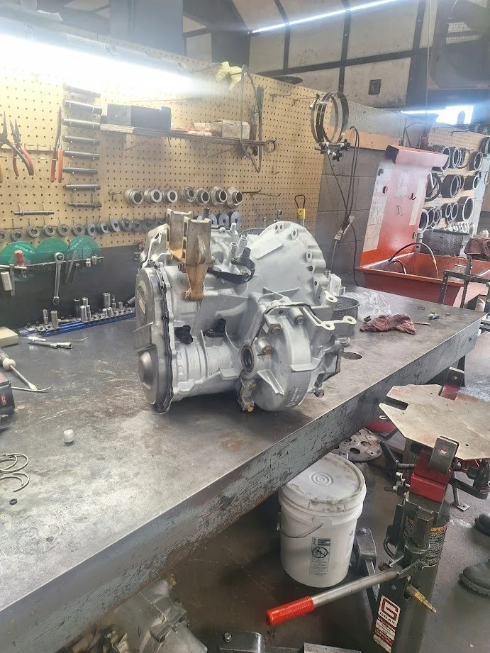 Work station with a mechanical part, likely a transmission, on a metal workbench in an industrial workshop with tools and parts mounted on pegboards in the background.