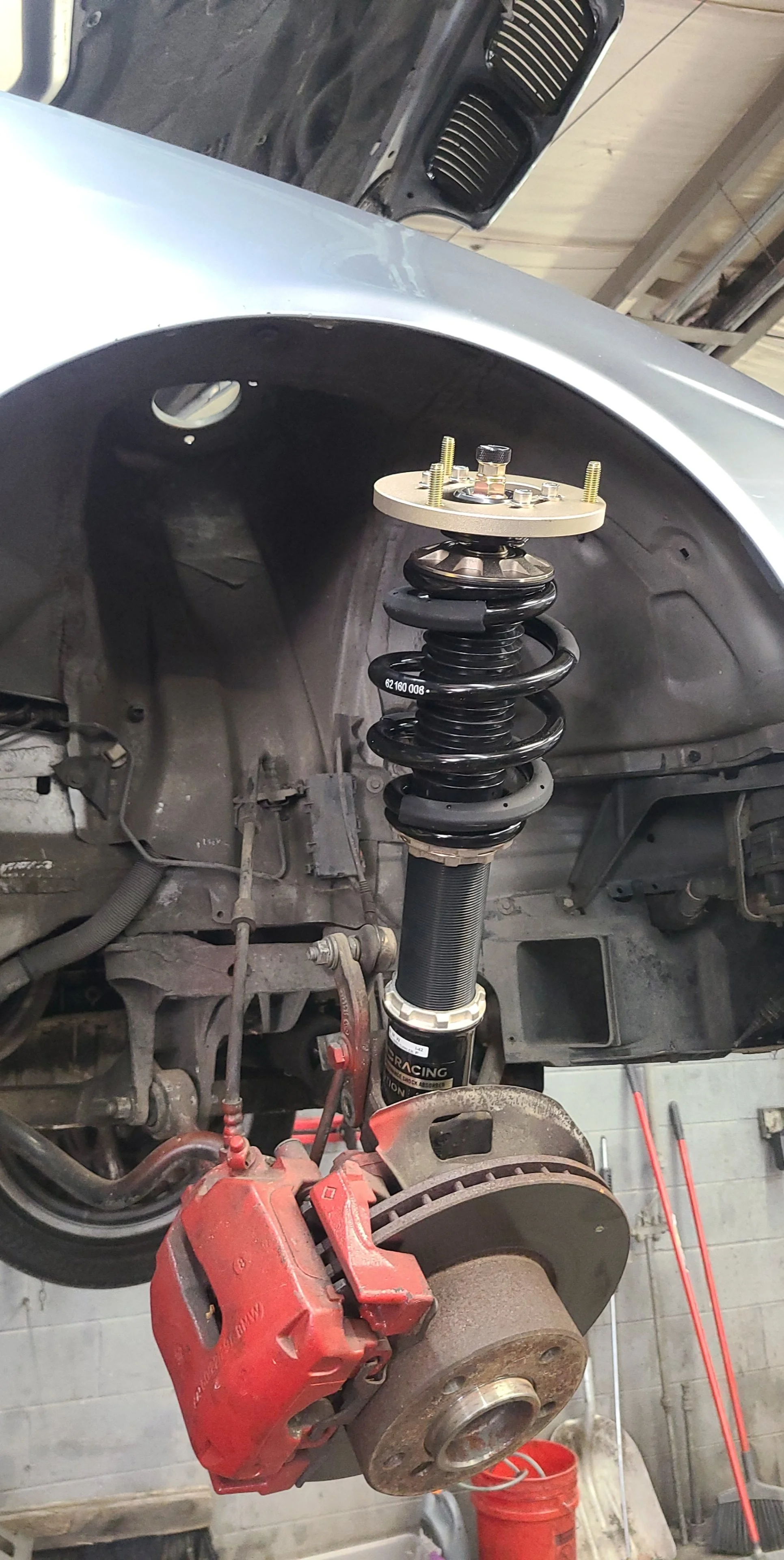 Close-up of a car suspension system with a strut assembly, coil spring, brake caliper, and rotor on a vehicle in a workshop.