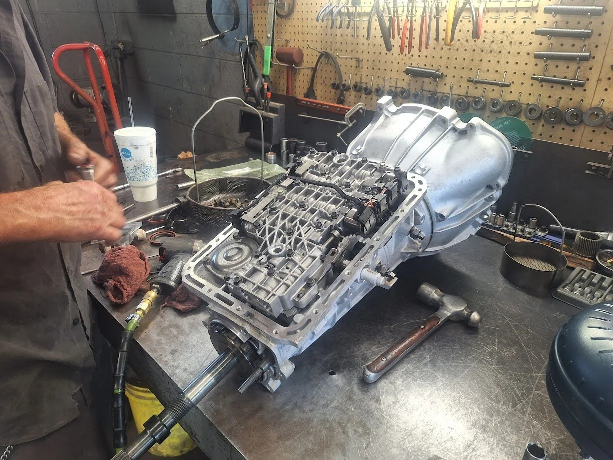 Automobile transmission disassembled on workbench with tools and parts in a garage workshop.