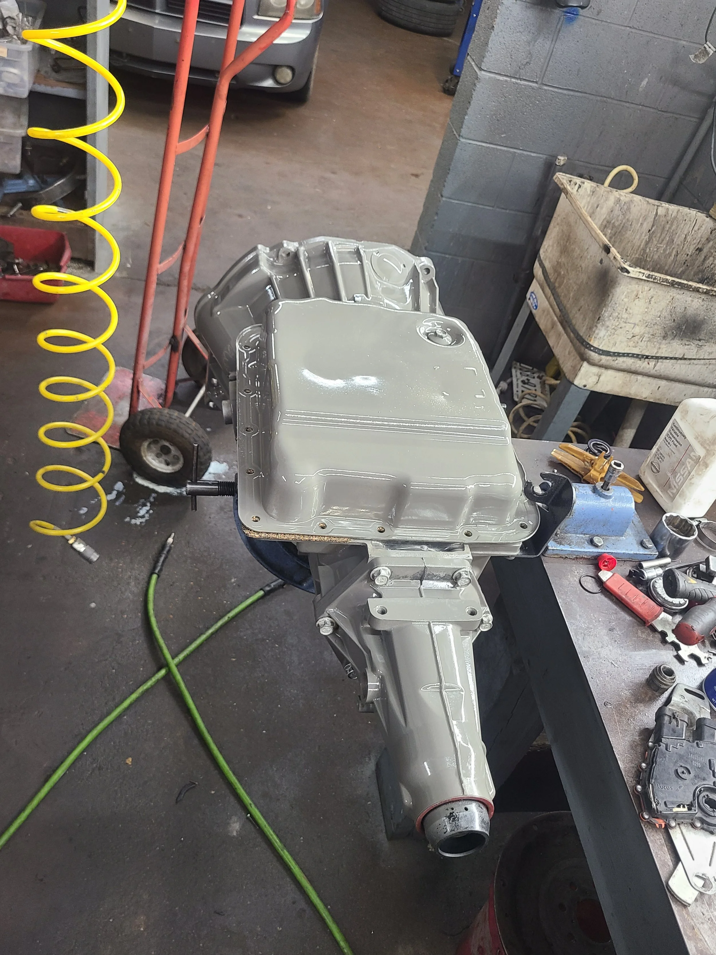 A partially assembled engine in a workshop, mounted on a black workbench with miscellaneous tools around, a concrete block wall, a tire, and a car in the background.