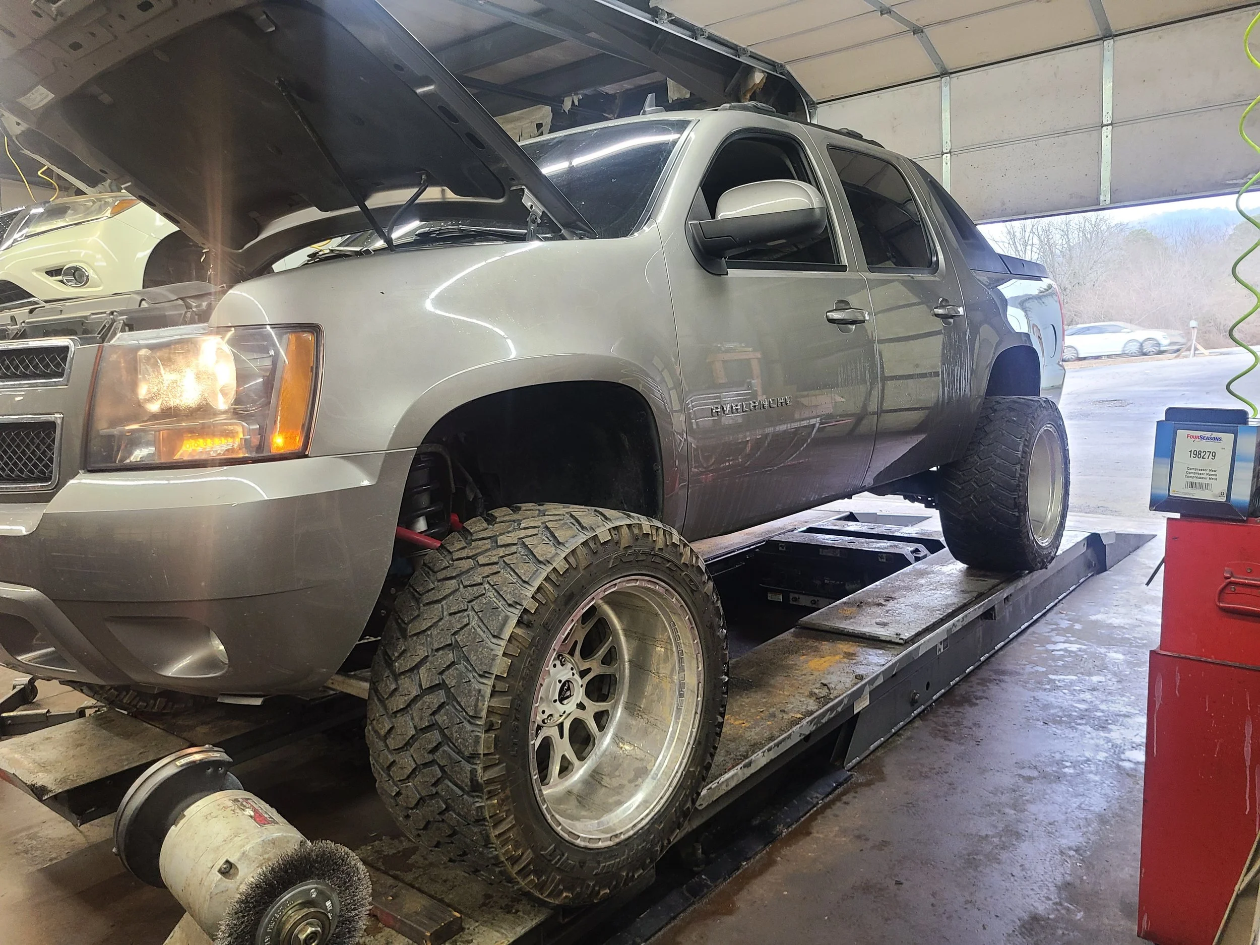 A gray Chevrolet Avalanche truck is on a dynamometer for testing, with the front left wheel removed and the hood open inside a garage.