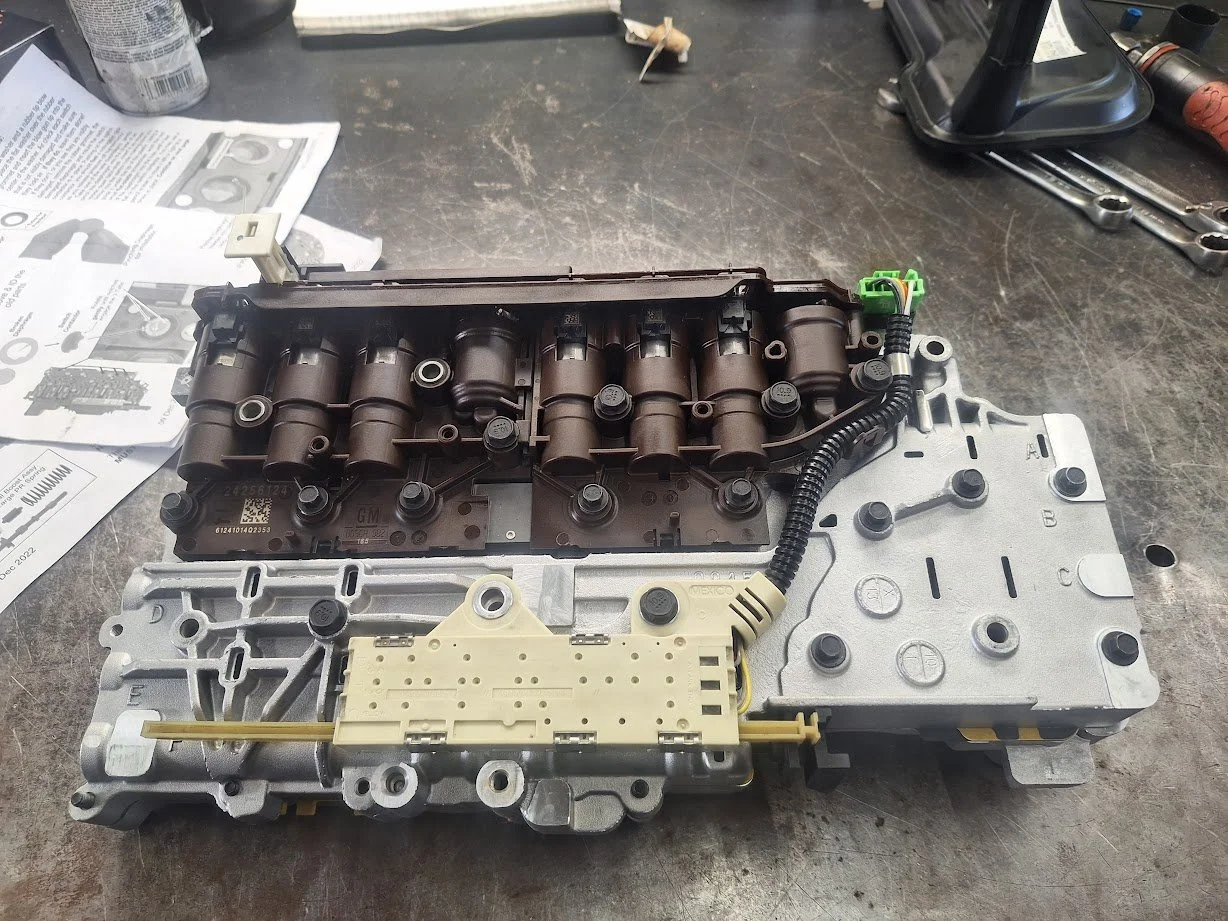 Automotive transmission valve body on workbench with tools and instructions nearby.
