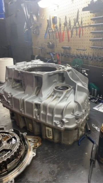 Automotive transmission housing on a workbench in a workshop with tools hanging on a pegboard in the background.