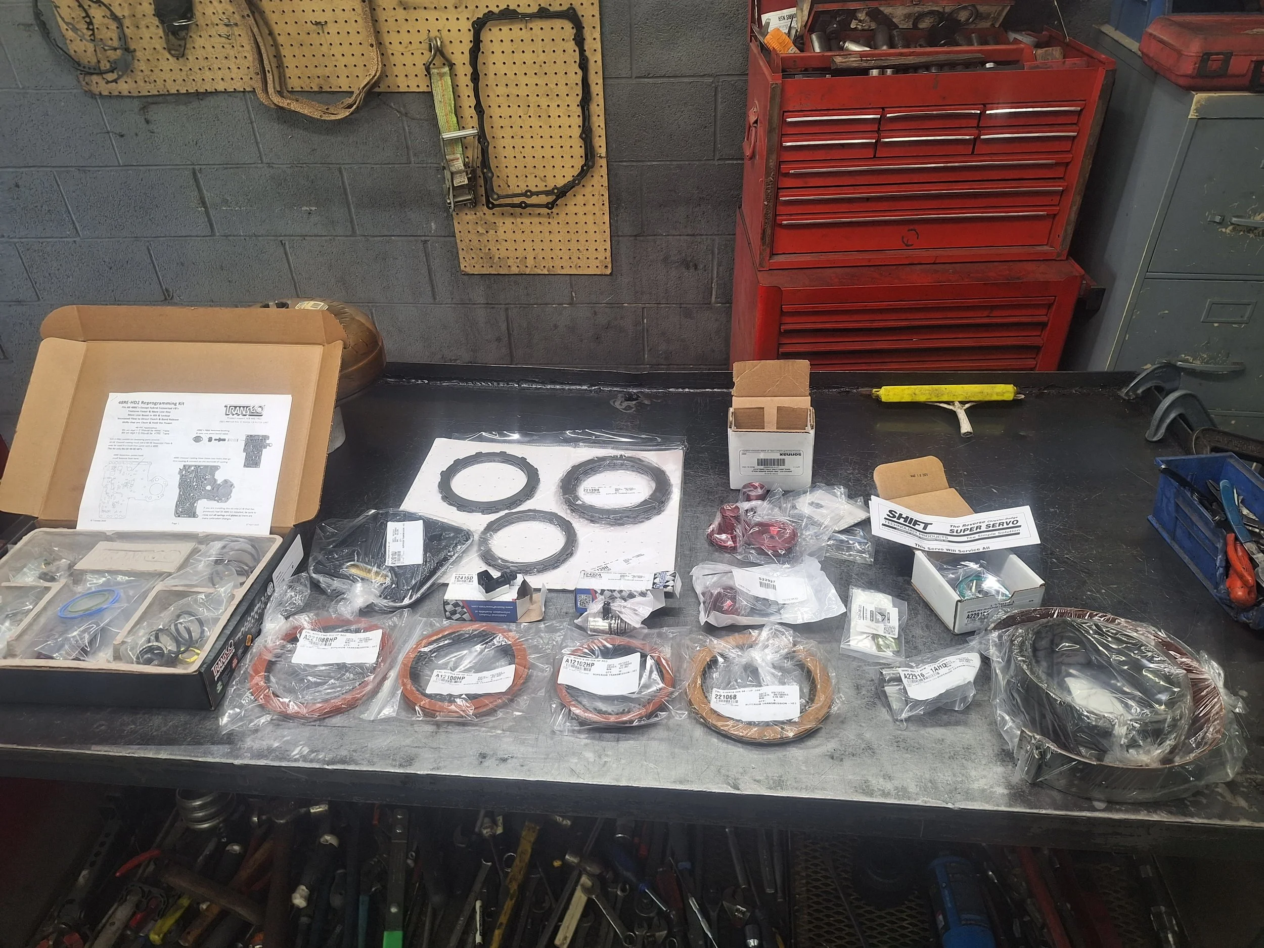 A workbench with assorted automotive parts and tools, including seals, O-rings, and packaged components, with a red tool chest and pegboard with tools in the background.