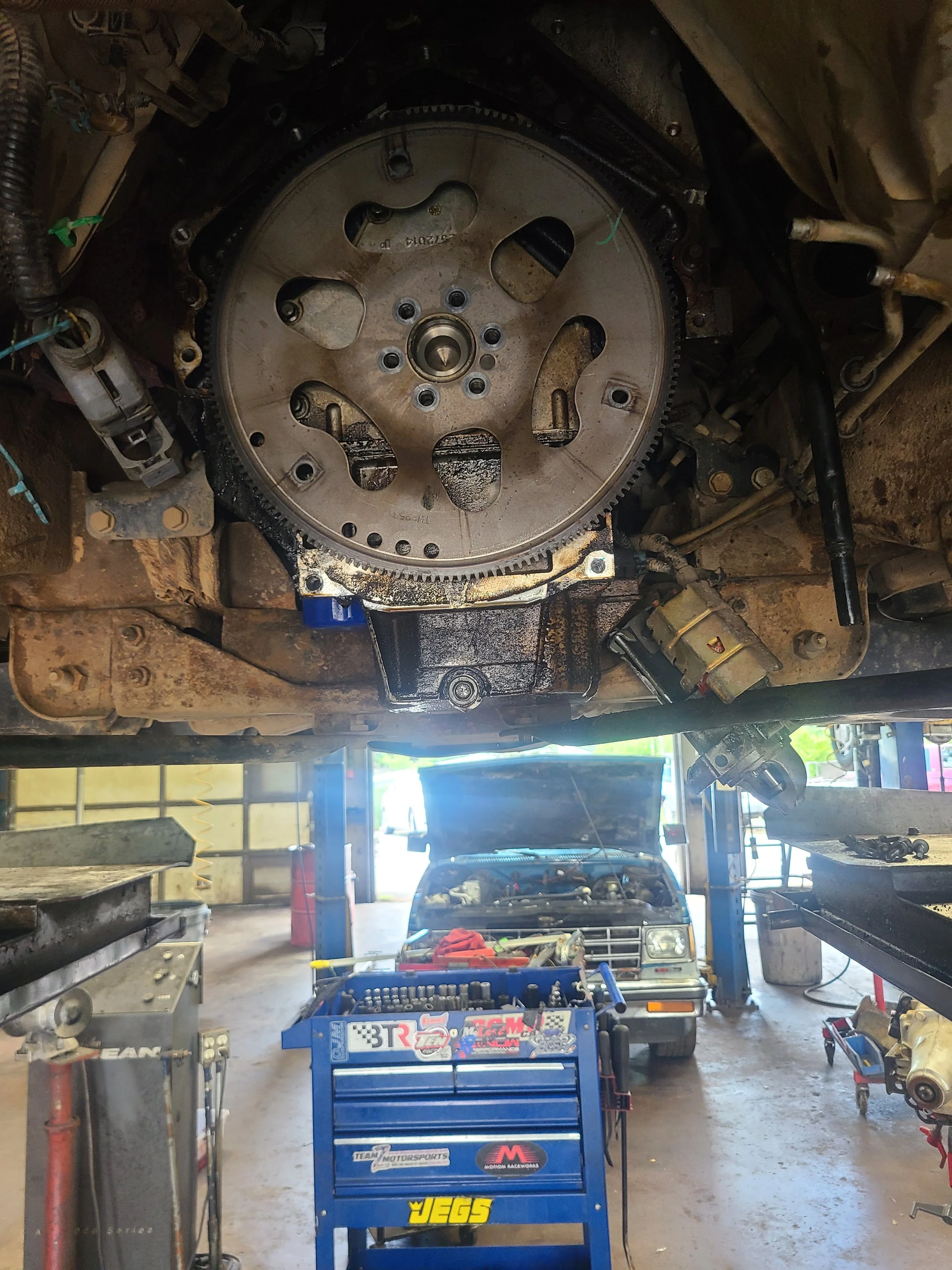 View of a car engine in a repair shop with the engine lifted and removed from the vehicle, exposing the flywheel. In the background, a vehicle with its hood open is parked inside the shop.