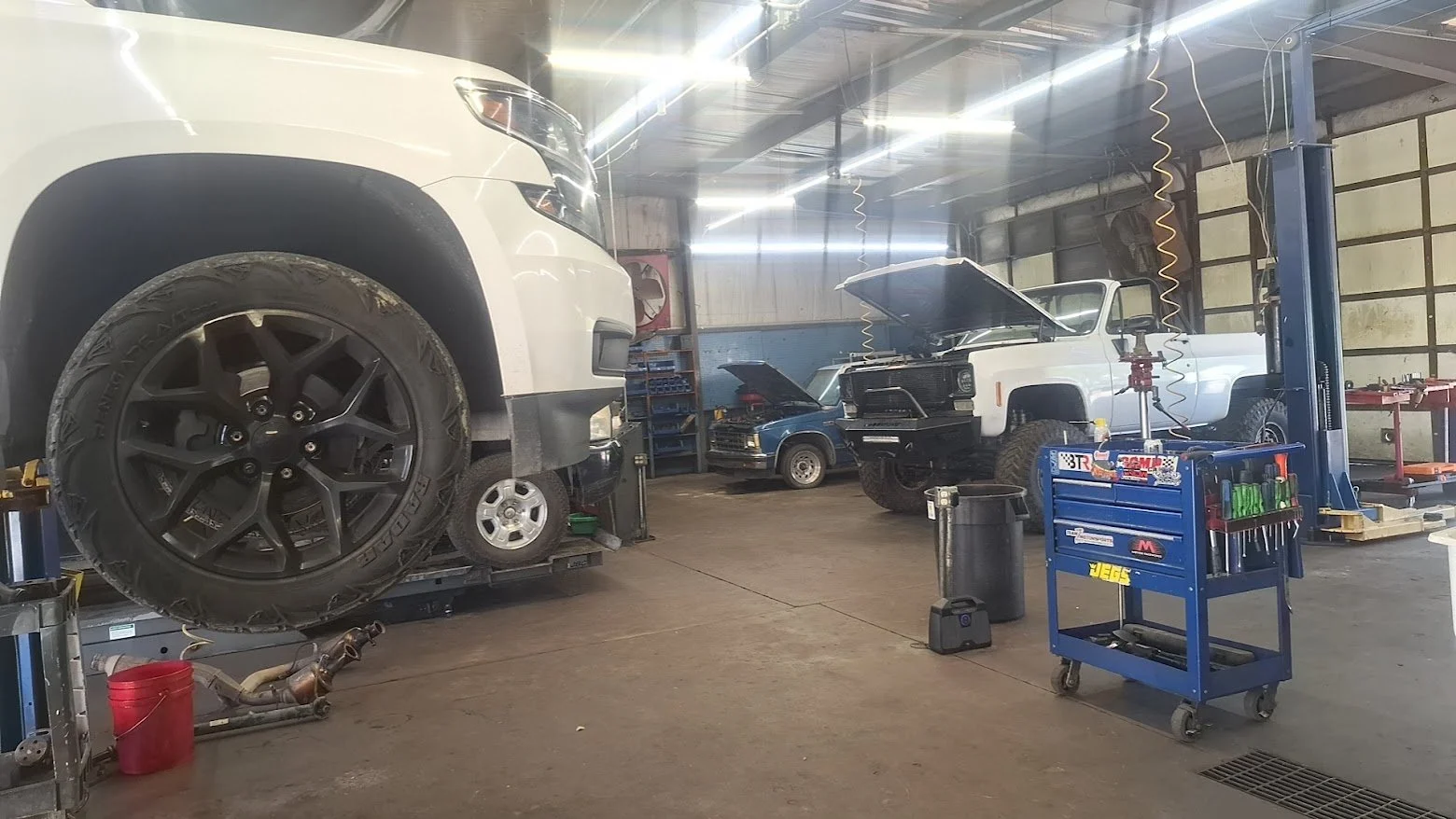 Inside an auto repair shop with vehicles in various stages of repair, including a white SUV on the lift, a vintage white pickup truck with an open hood, and a classic blue car. Tools and equipment are visible, including a tool chest and carts.