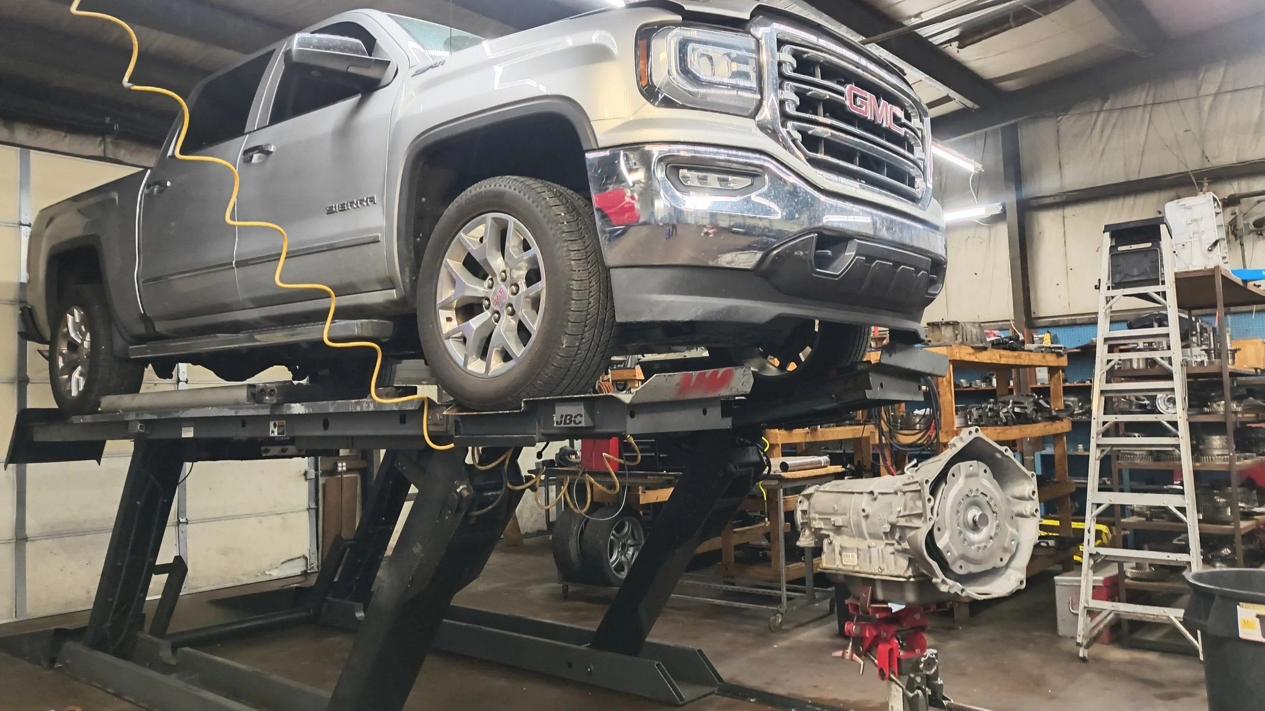GMC pickup truck elevated on a hydraulic lift inside an auto repair shop, with a transmission on the ground nearby, shelves with tools, ladder, and workshop equipment in the background.