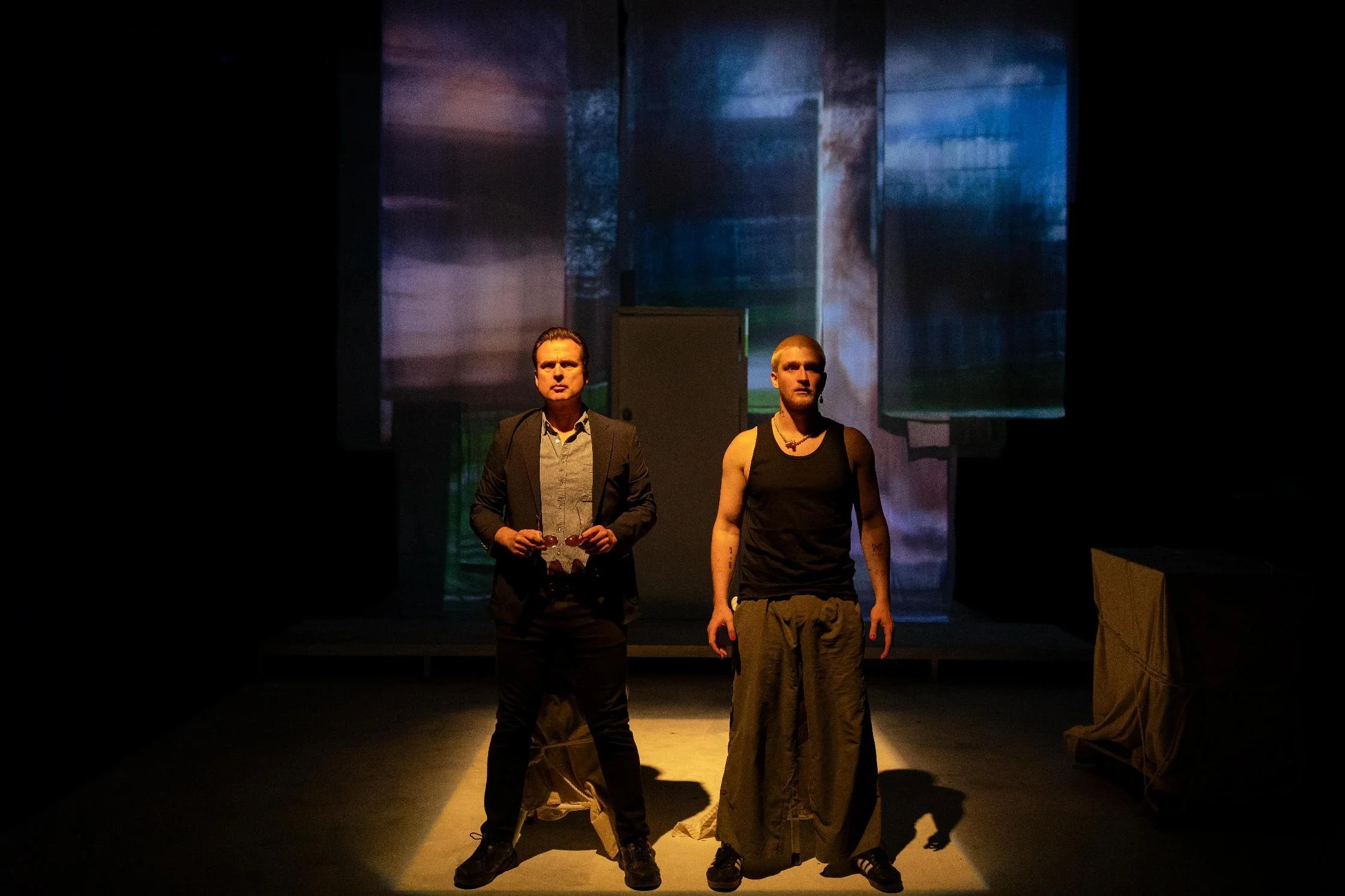 Two men stand on a dimly lit stage, with one wearing a suit and holding glasses, and the other in casual clothing with tattoos, in front of a colorful abstract background.