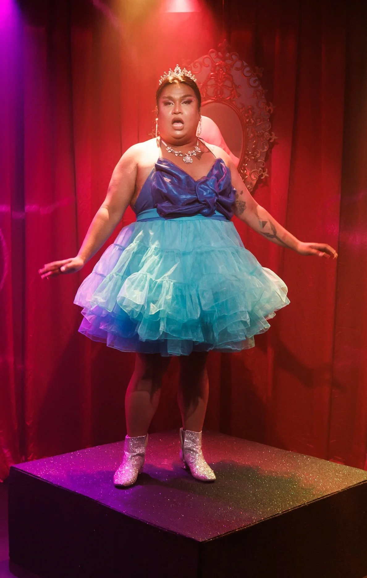 A person standing on a glittery platform, dressed in a colorful, tutu-style dress with a sparkly top, wearing glittery boots, jewelry, and a tiara, with a red curtain background and ornate mirror.