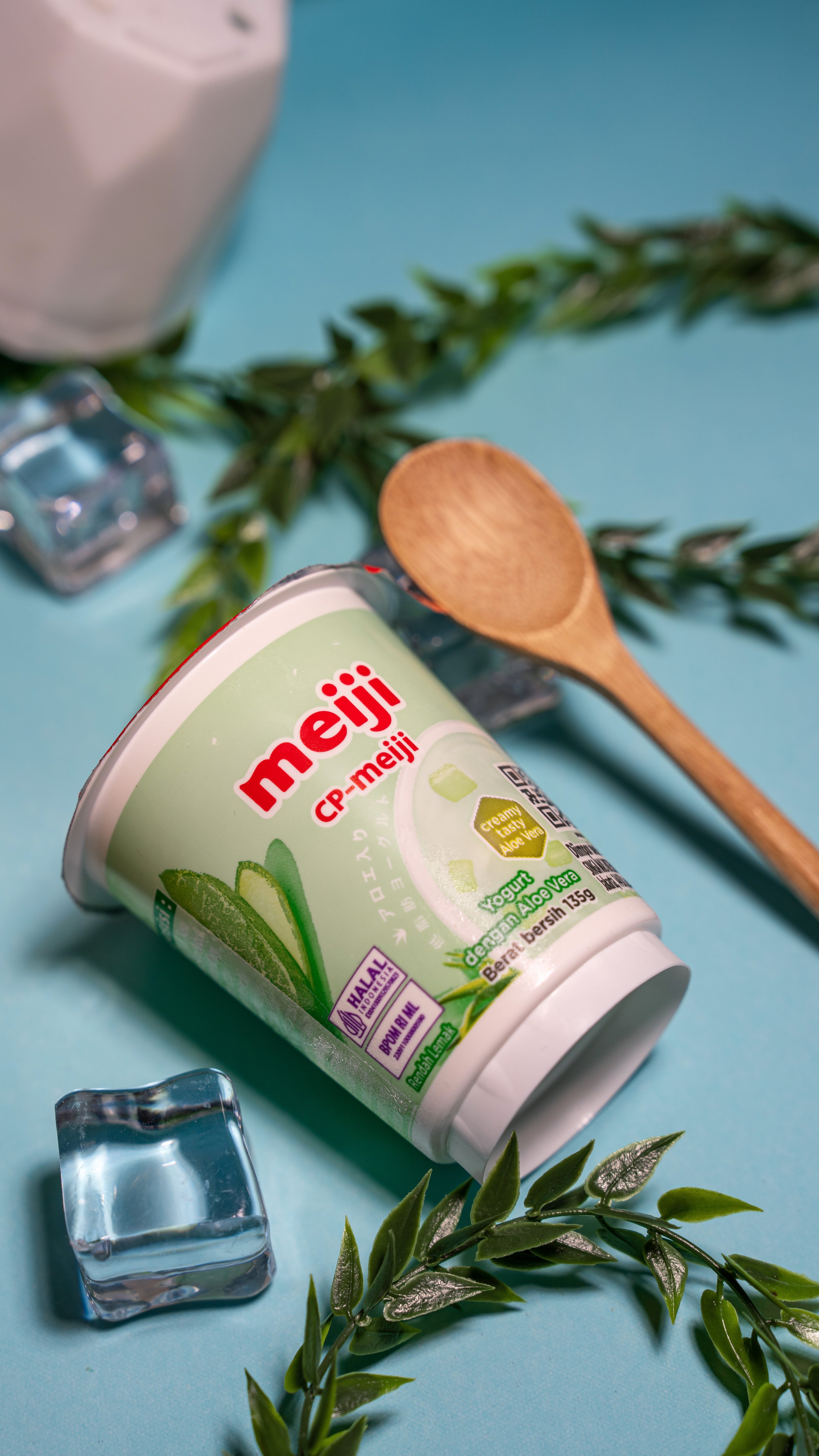 A cup of Meiji CP-meiji creamy aloe vera yogurt surrounded by green leaves, ice cubes, and a wooden spoon on a blue background.