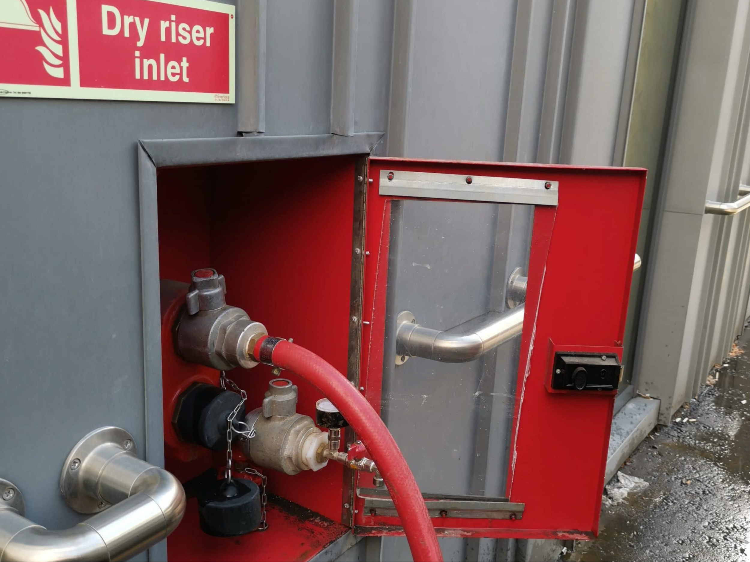 Industrial fire hydrant inside a bright red cabinet with a transparent door, attached to a gray wall, with a hose connected to it, near a sign that reads 'Dry riser inlet'.