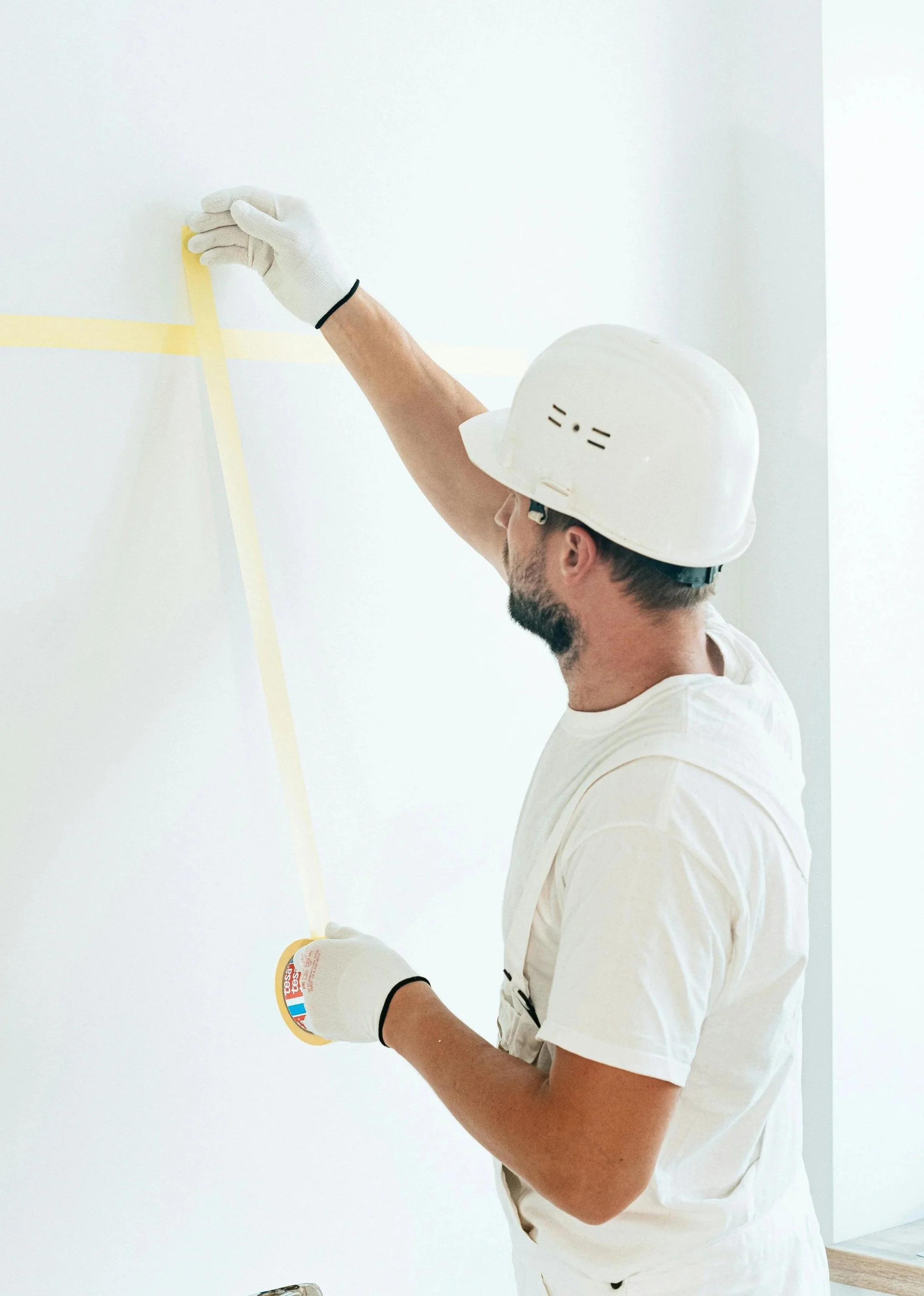 A man wearing a white work helmet and white gloves is taping painter's tape on a white wall.