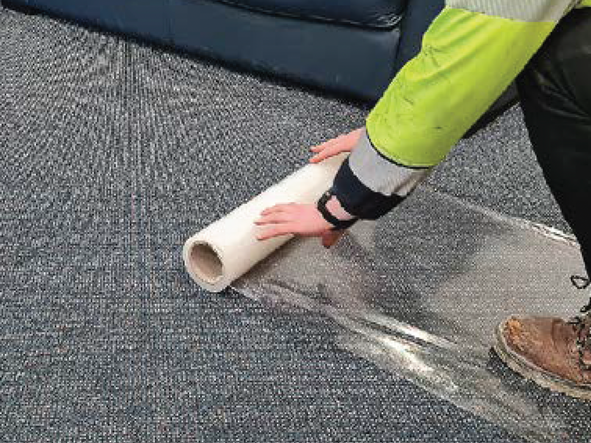 Person in a green and gray jacket unrolling a carpet on a gray textured surface.