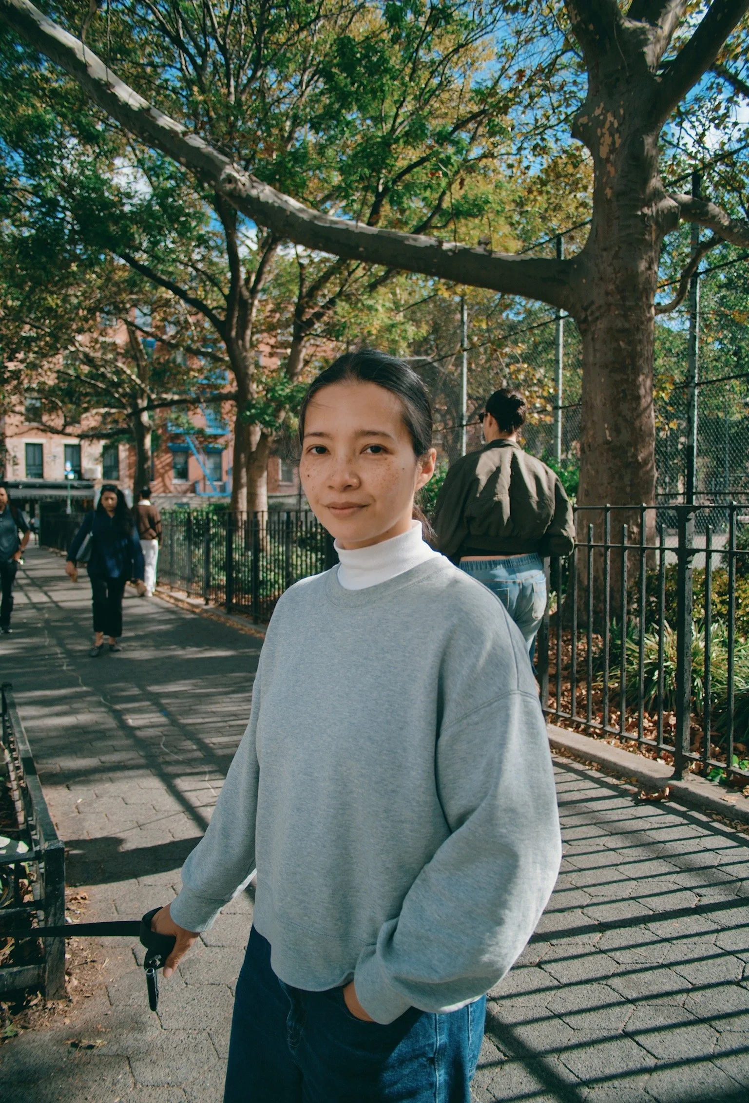 A young woman with dark hair, wearing a gray sweatshirt and a white turtleneck, standing on a sidewalk in a park during autumn, holding a set of car keys, with trees and other people in the background.