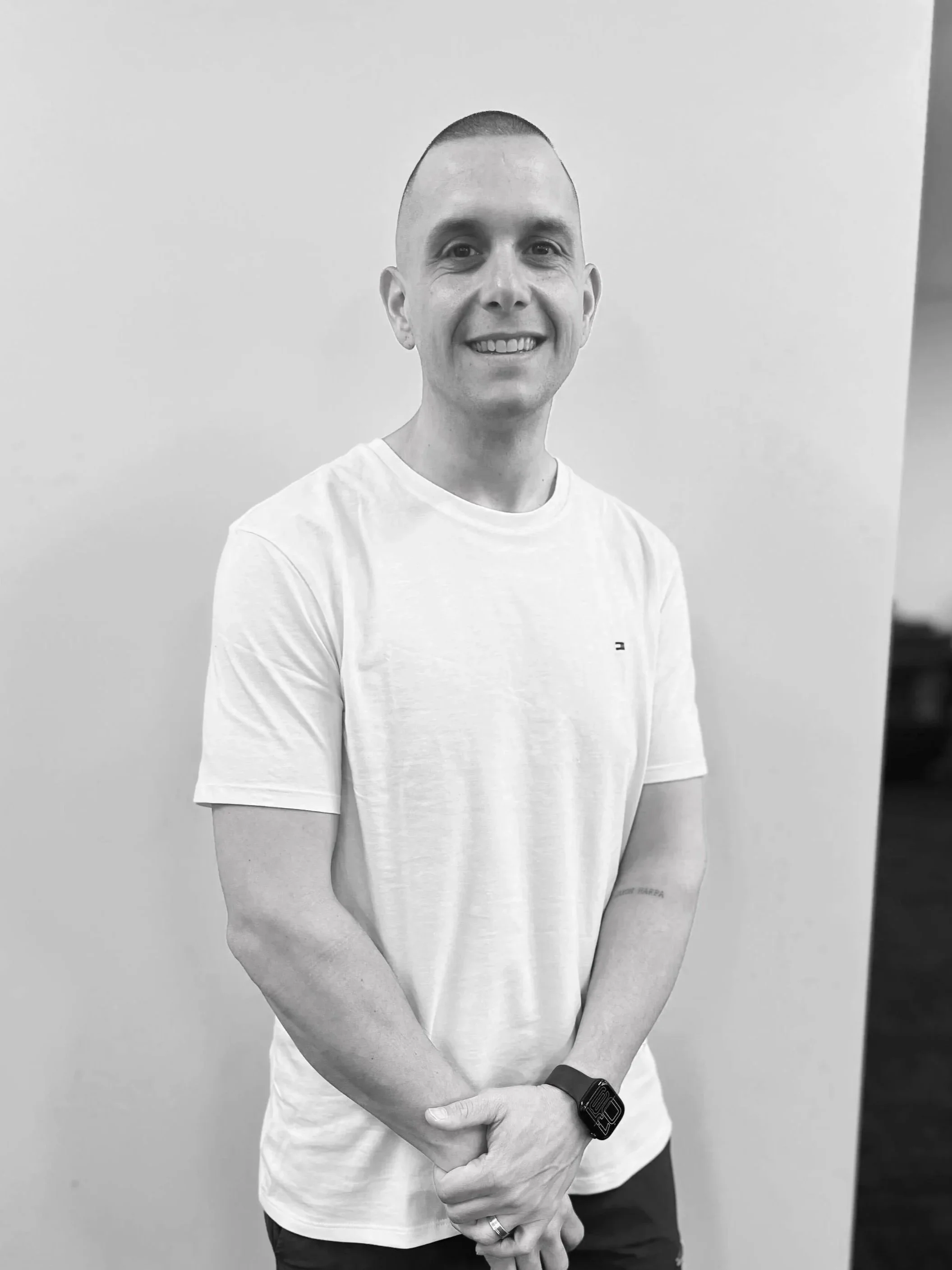 A smiling man with a buzz cut, wearing a white t-shirt, standing against a plain wall.