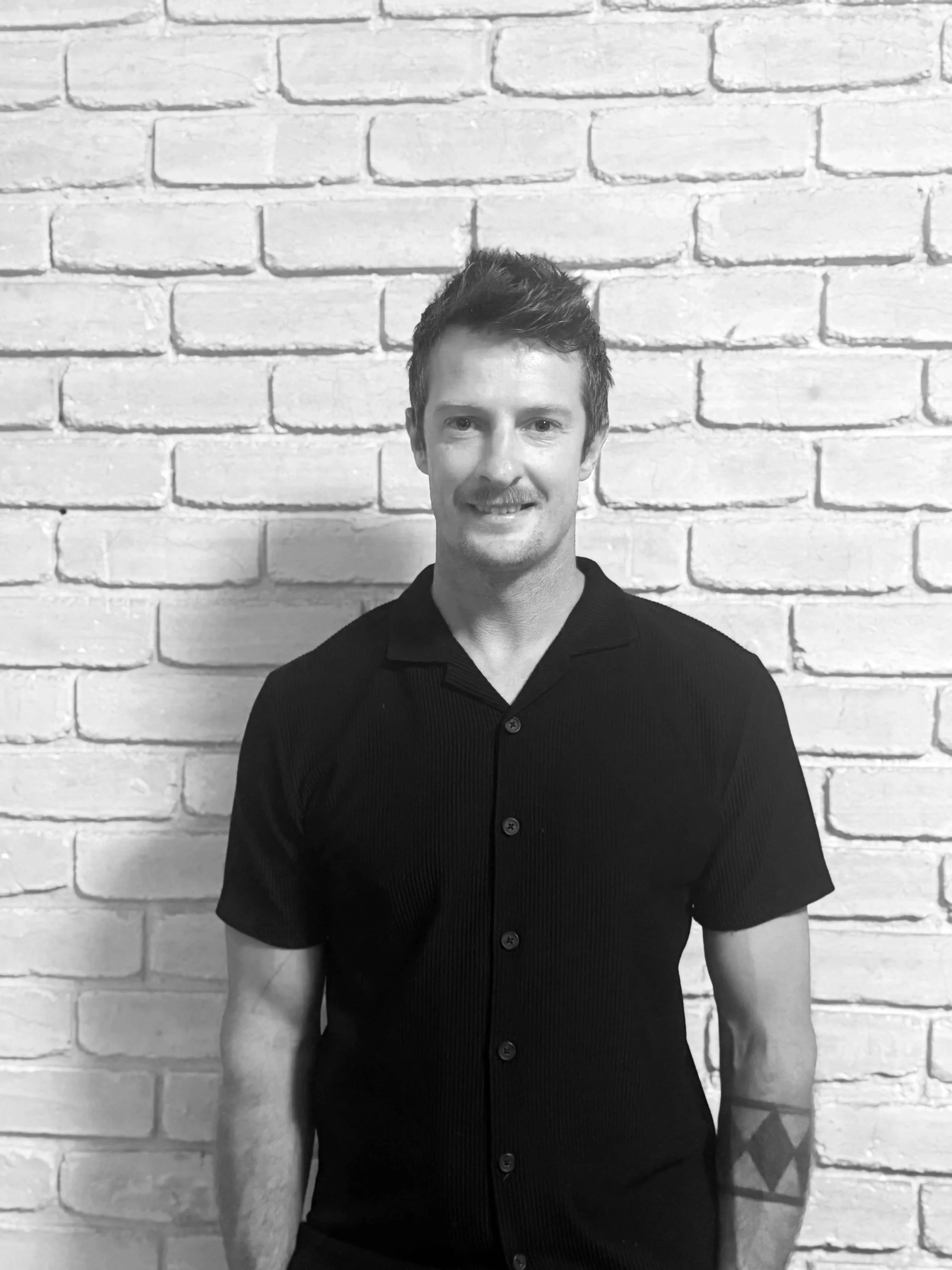 Black and white portrait of a smiling man with short, styled hair and a mustache, standing against a brick wall, wearing a short-sleeved, button-up shirt.