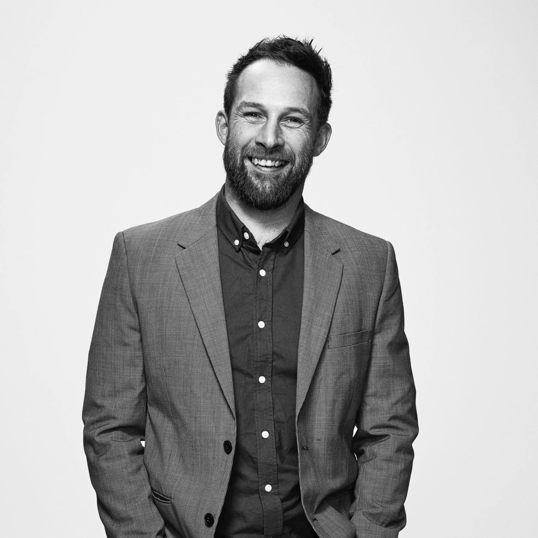 Black and white portrait of a smiling man with a beard, wearing a suit jacket and button-up shirt, standing against a plain background.