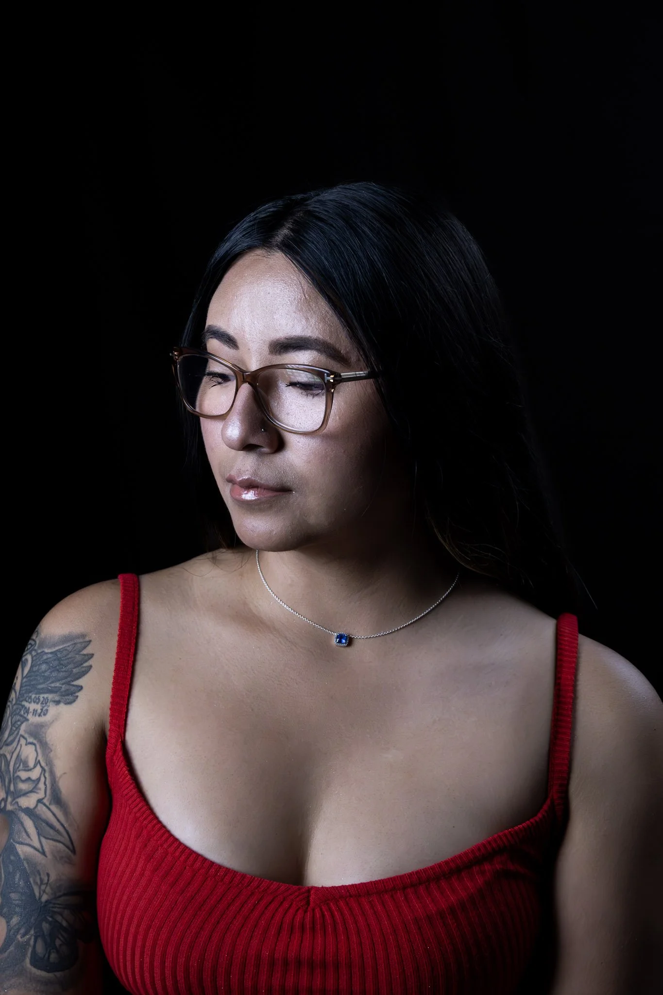 A woman with long dark hair wearing glasses, a red sleeveless top, and a silver necklace with a blue gemstone, looking down against a black background.