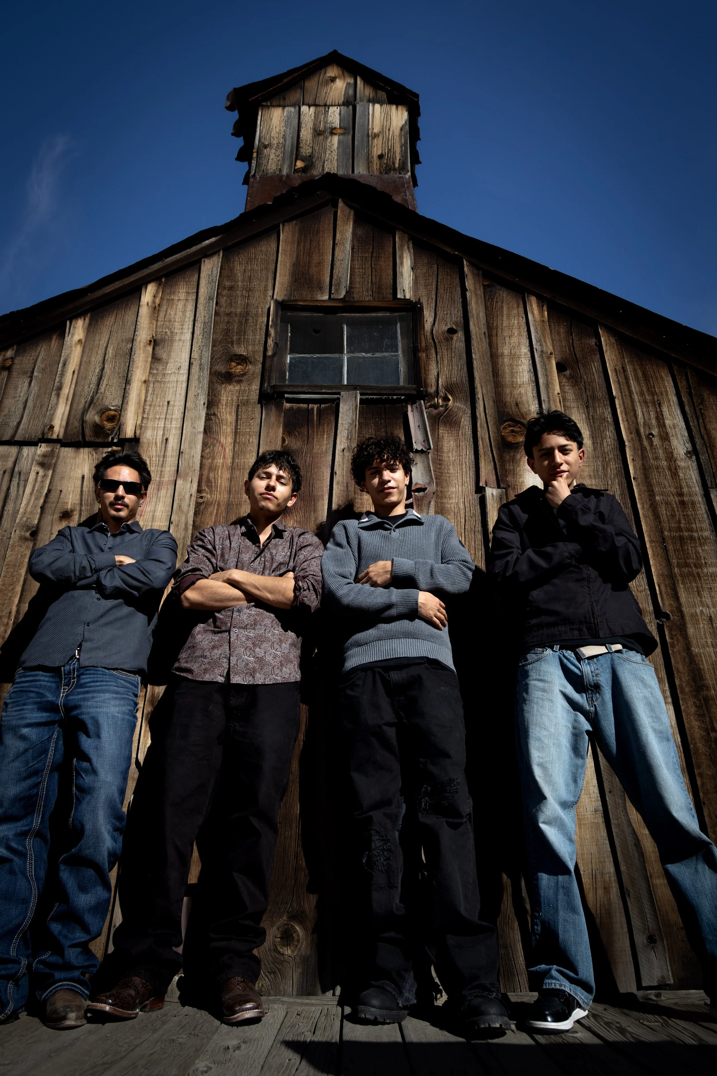 Four young men standing in front of a wooden building with a small tower on top, posing confidently with arms crossed or hands near their faces, under a clear blue sky.