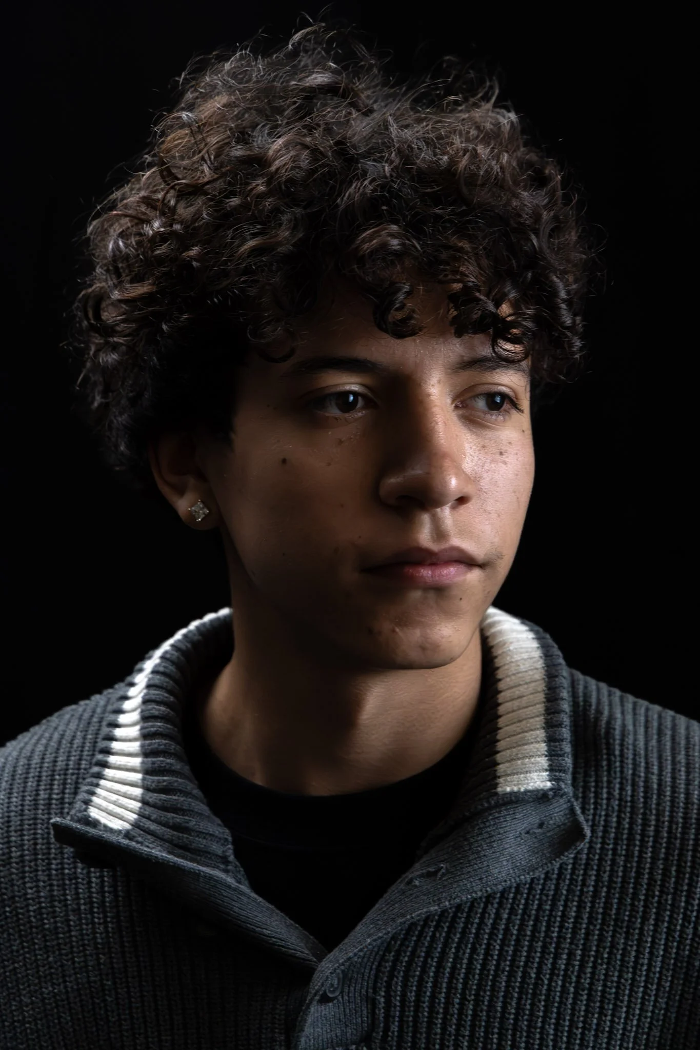 Portrait of a young woman with curly hair, wearing a dark striped sweater, and a black background.