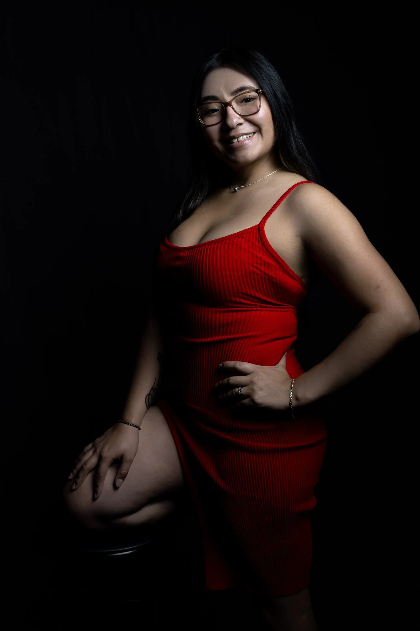 A woman with glasses and long dark hair wears a red dress and poses against a black background.
