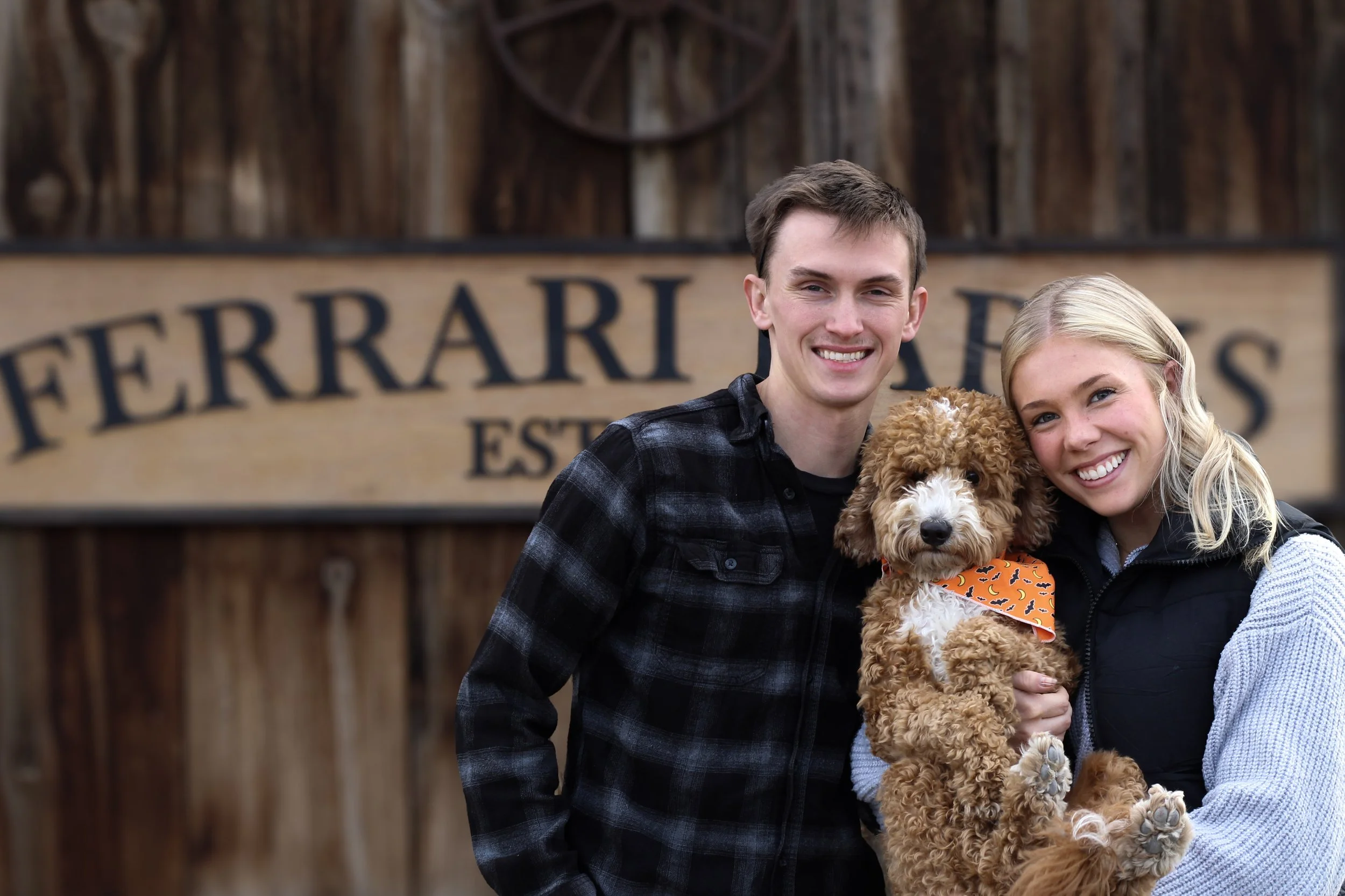 A young couple smiling, holding a curly-haired dog with an orange bandana, standing in front of a rustic wooden sign that reads "Ferrari".